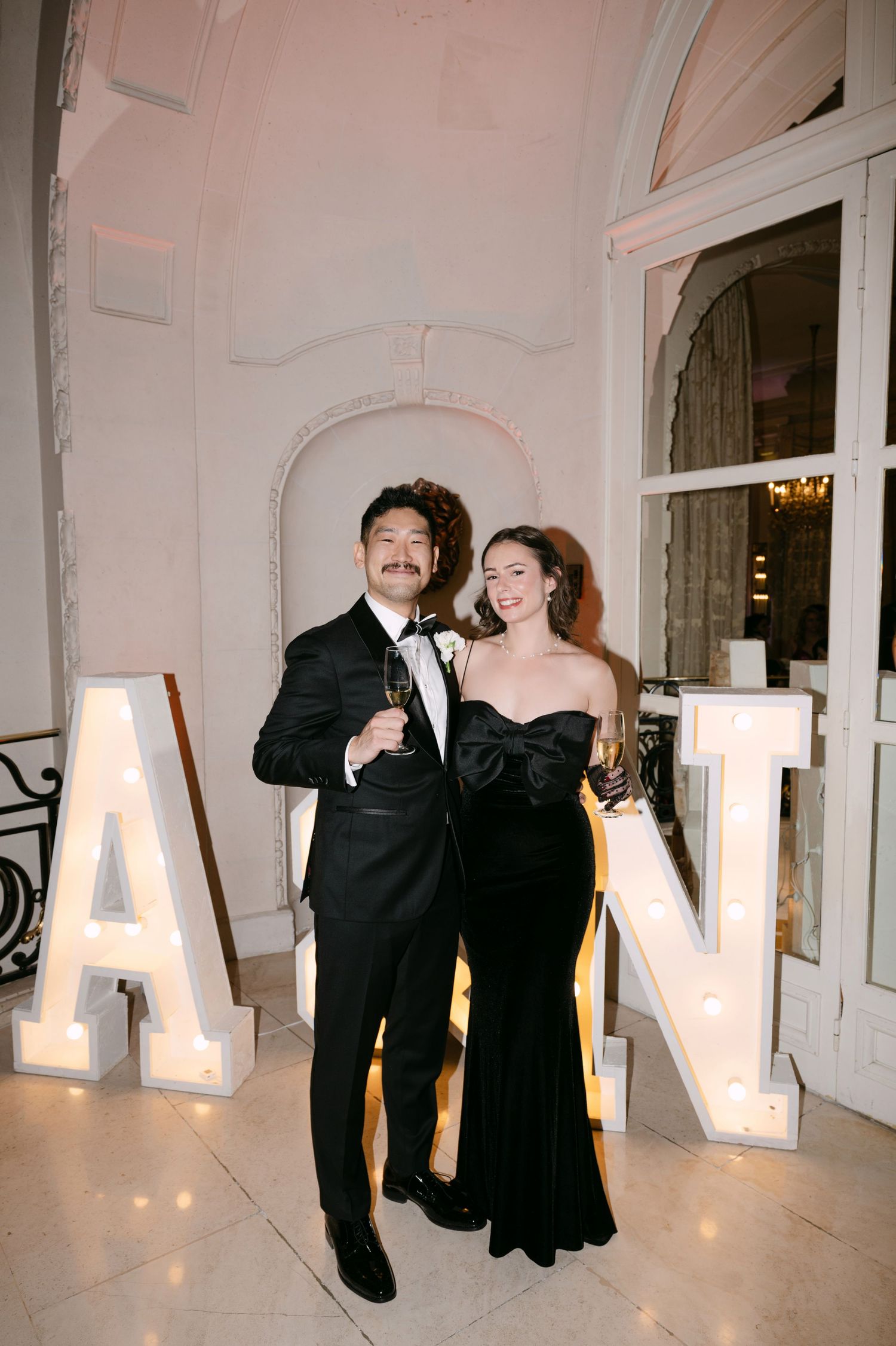 Couple in black formal attire poses smiling between illuminated A and N marquee letters at wedding reception venue.