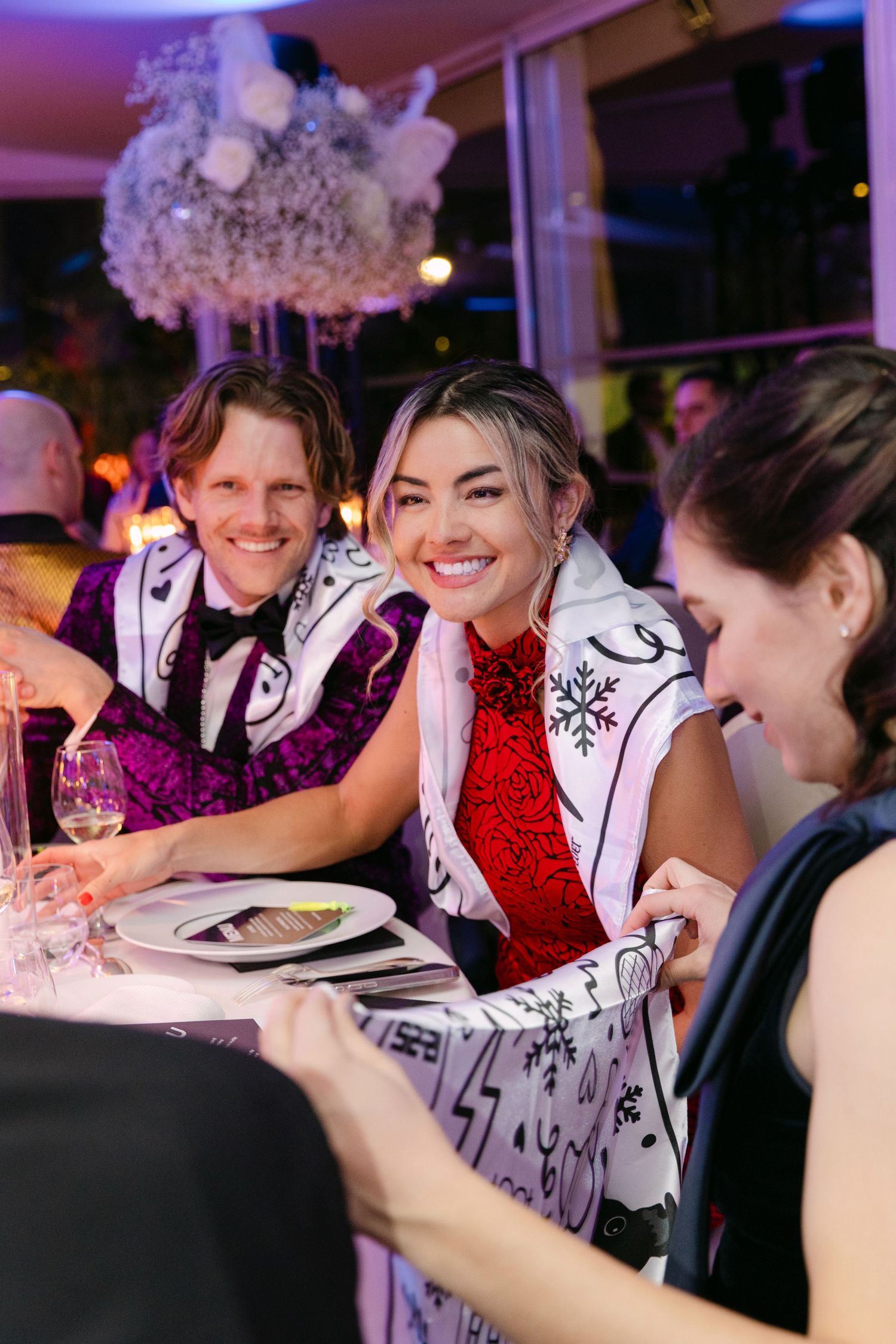 Smiling wedding guests wrapped in festive scarves seated at an elegant dinner table at a winter gala event.