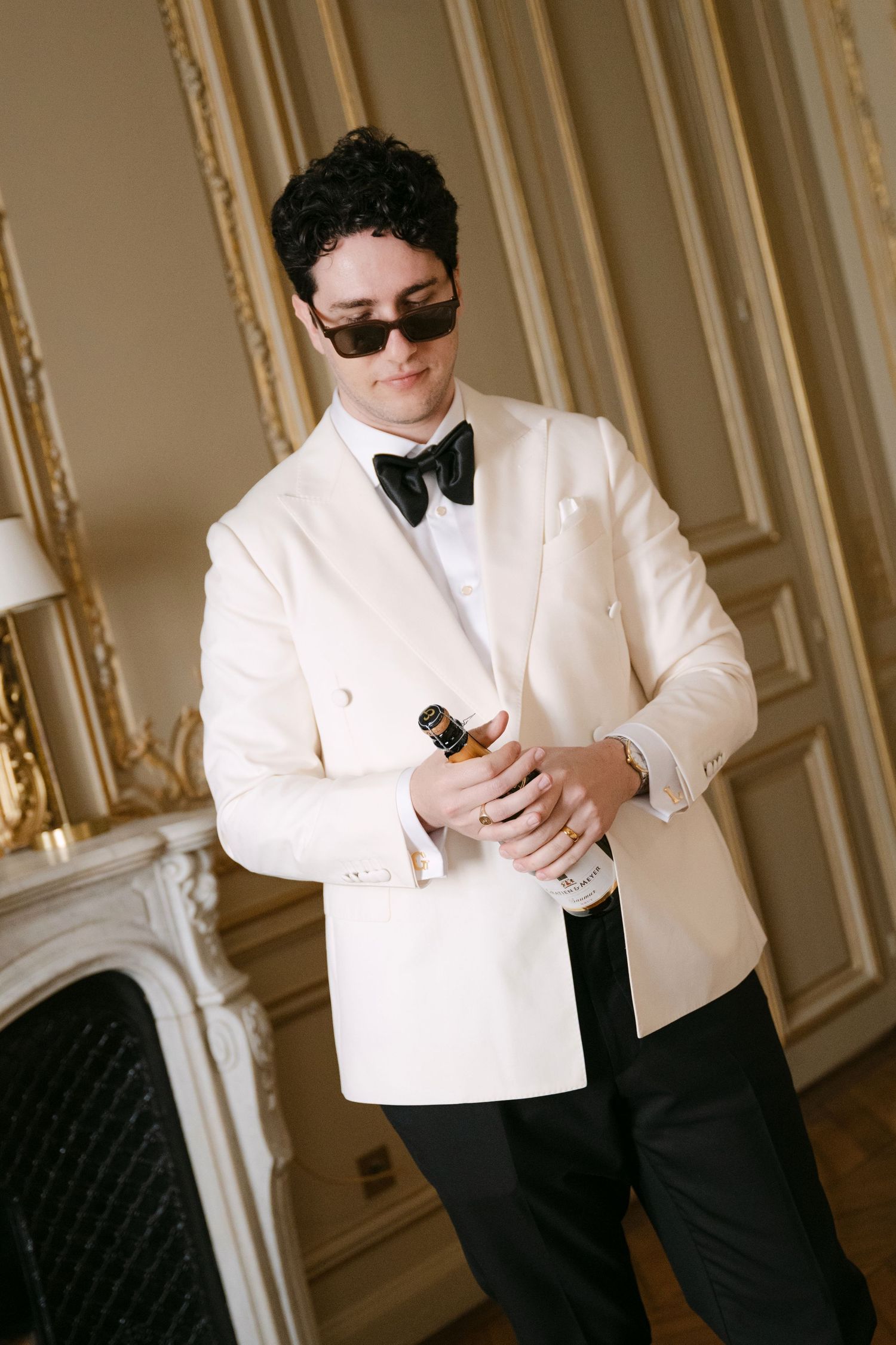 A person in a formal white tuxedo jacket and black pants adjusts their outfit in an elegant room with ornate walls.