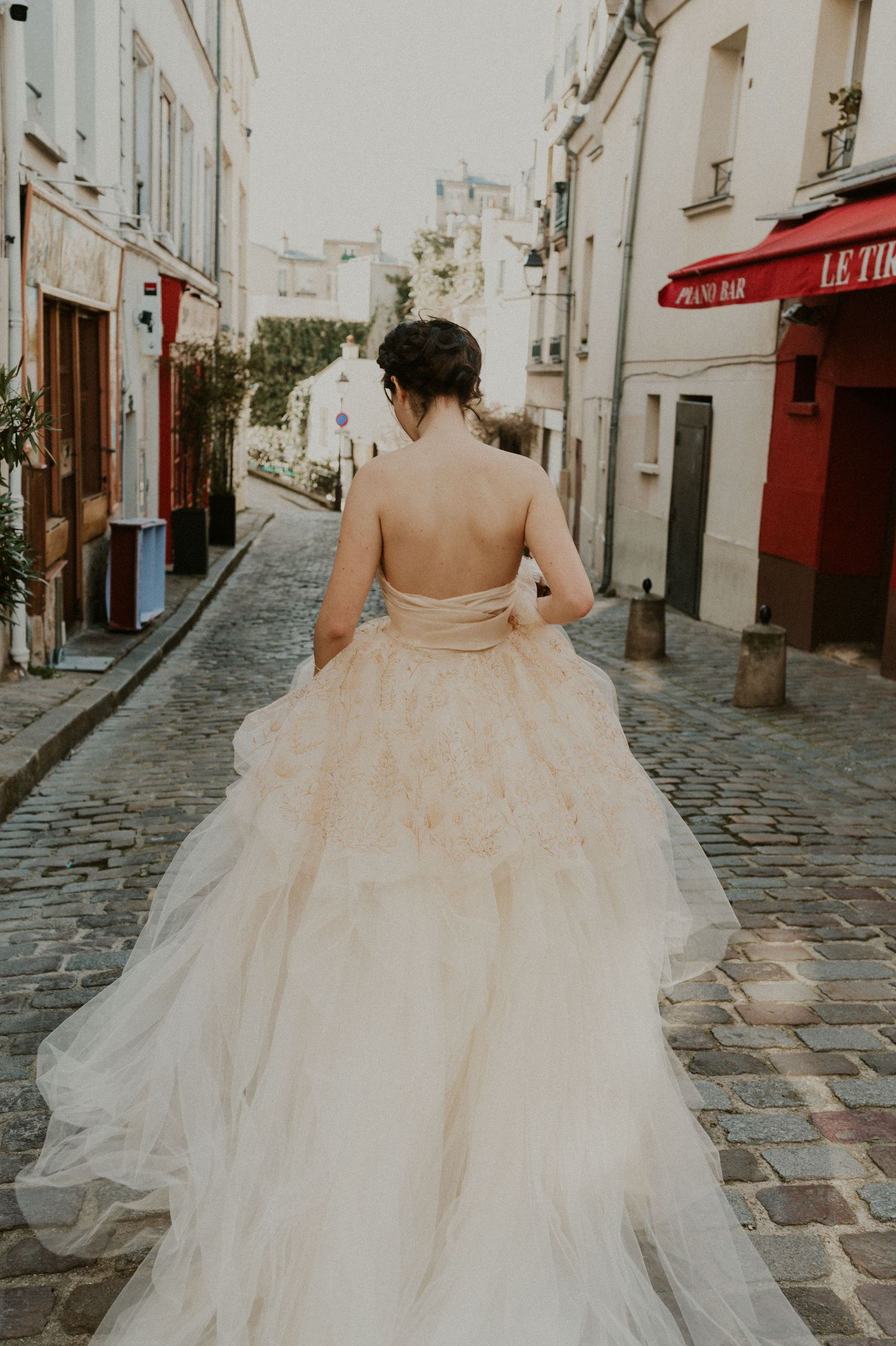 Back view of bride in flowing champagne gown walking down historic cobblestone street.