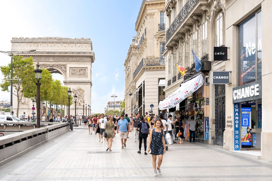 Arc de Triomphe visible at end of shopping street with tourists and storefronts.