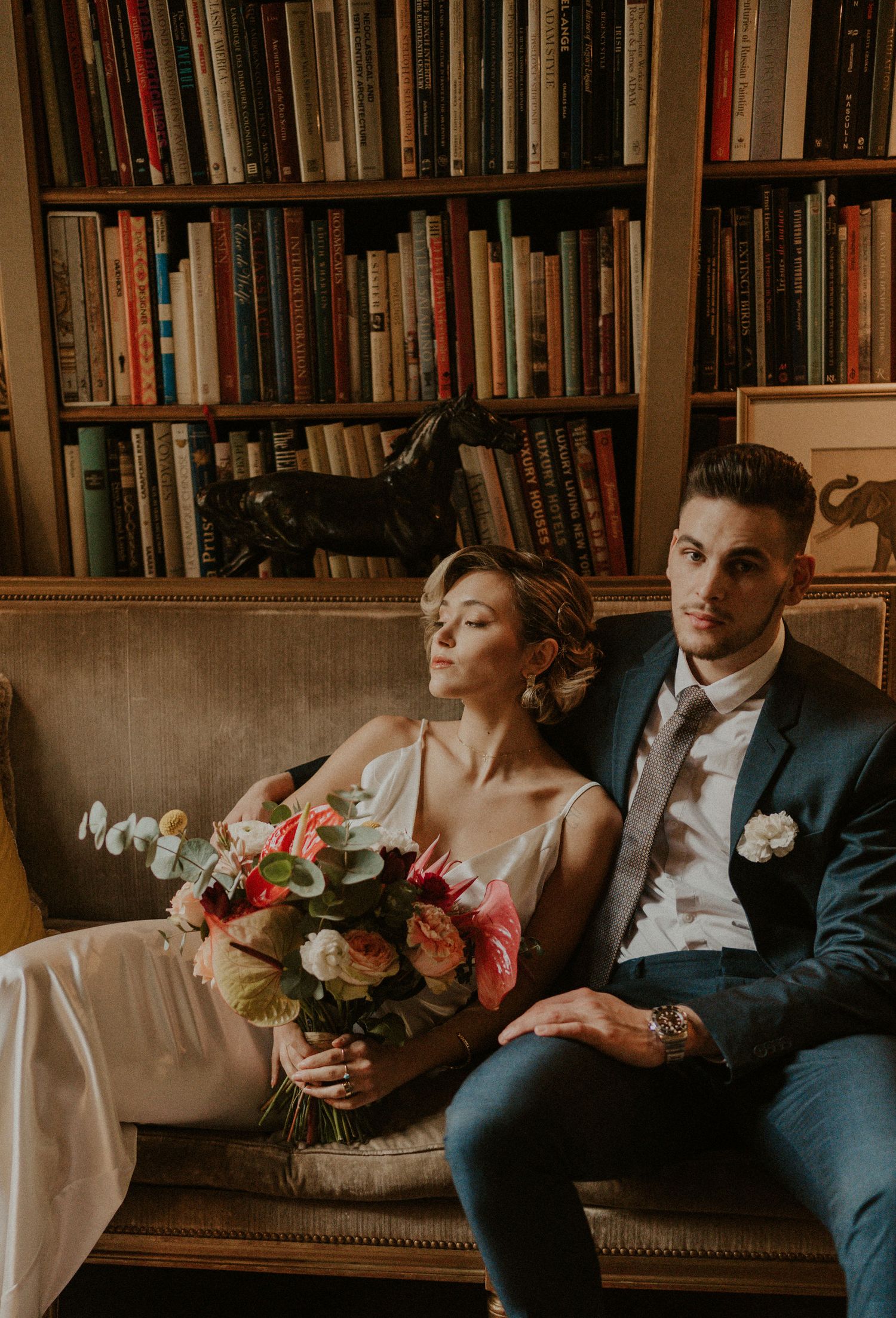 Elegant couple in wedding attire relaxes on vintage couch with floral bouquet against library backdrop.
