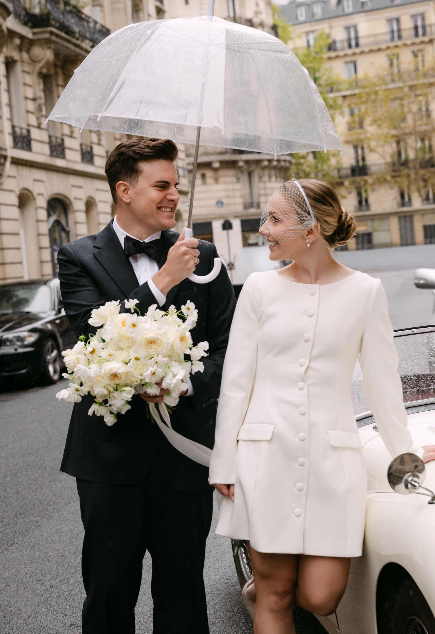 A romantic wedding couple shares joyful moments on a Parisian street wearing elegant white and black attire.