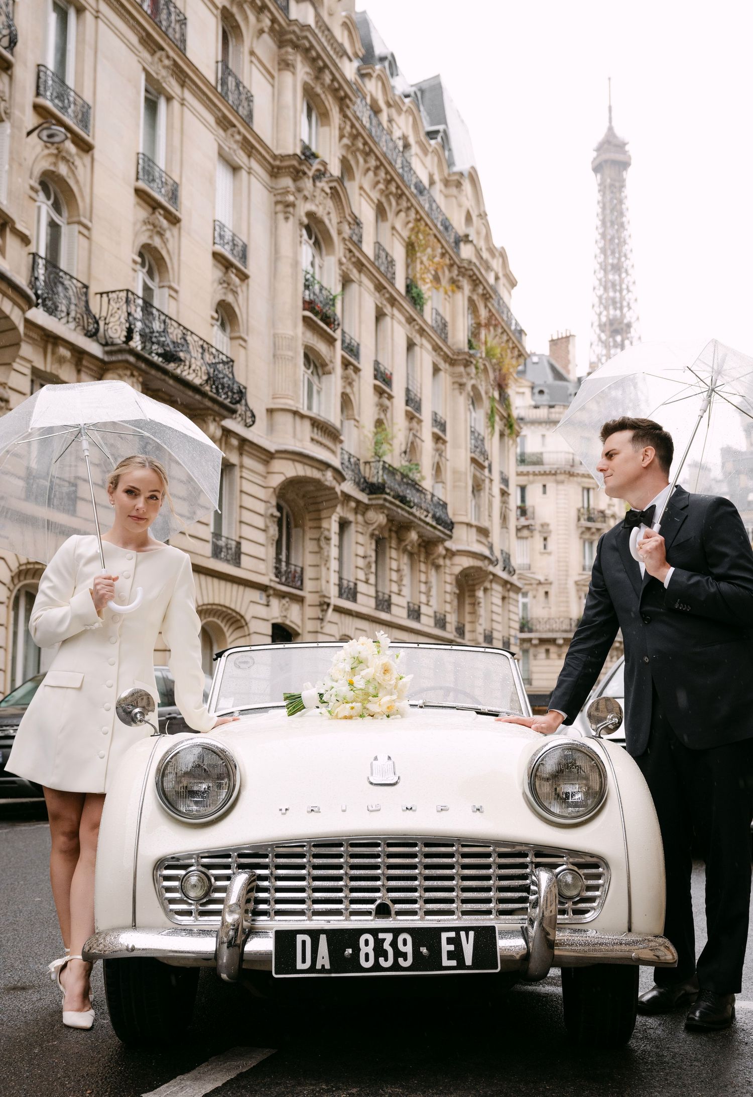 Vintage white Triumph car parked on a Paris street with the Eiffel Tower visible in the background on a rainy day.