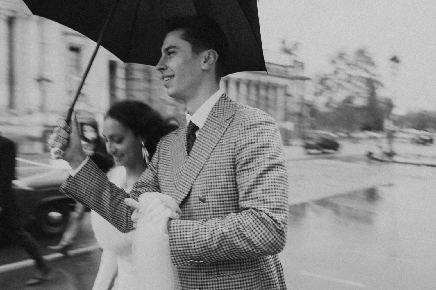 Black and white candid moment of a couple with umbrella on a rainy city street.
