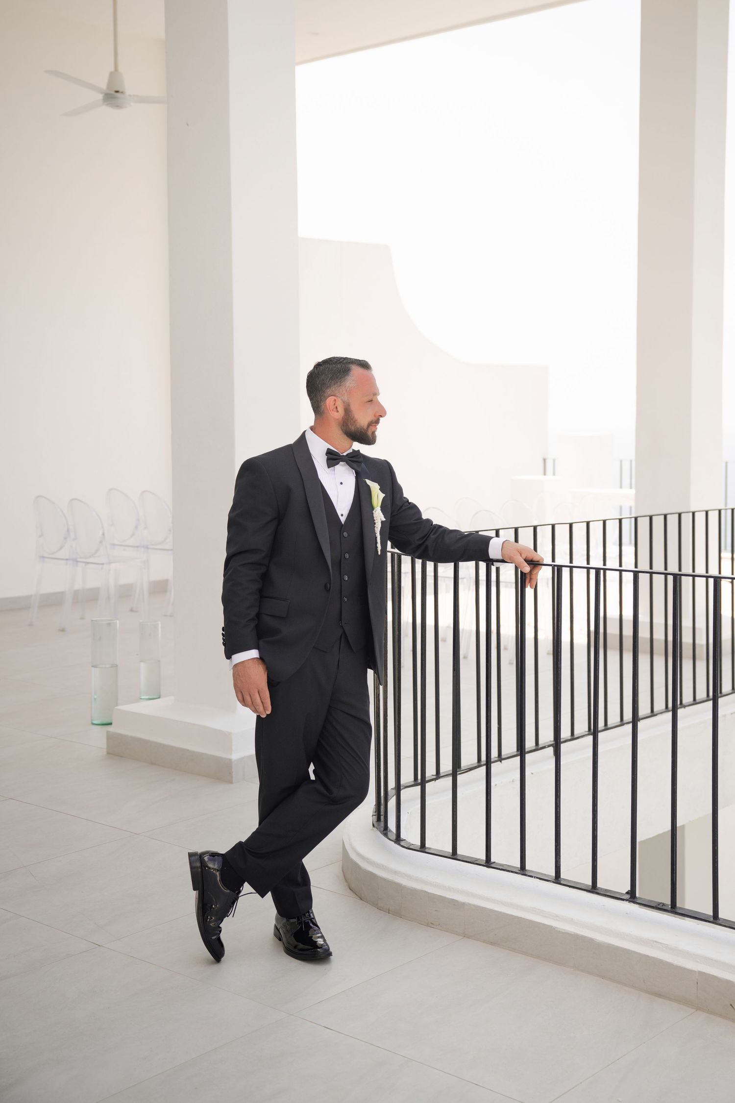 Groom in black tuxedo with bow tie leans against iron railing in bright white venue, looking away confidently.