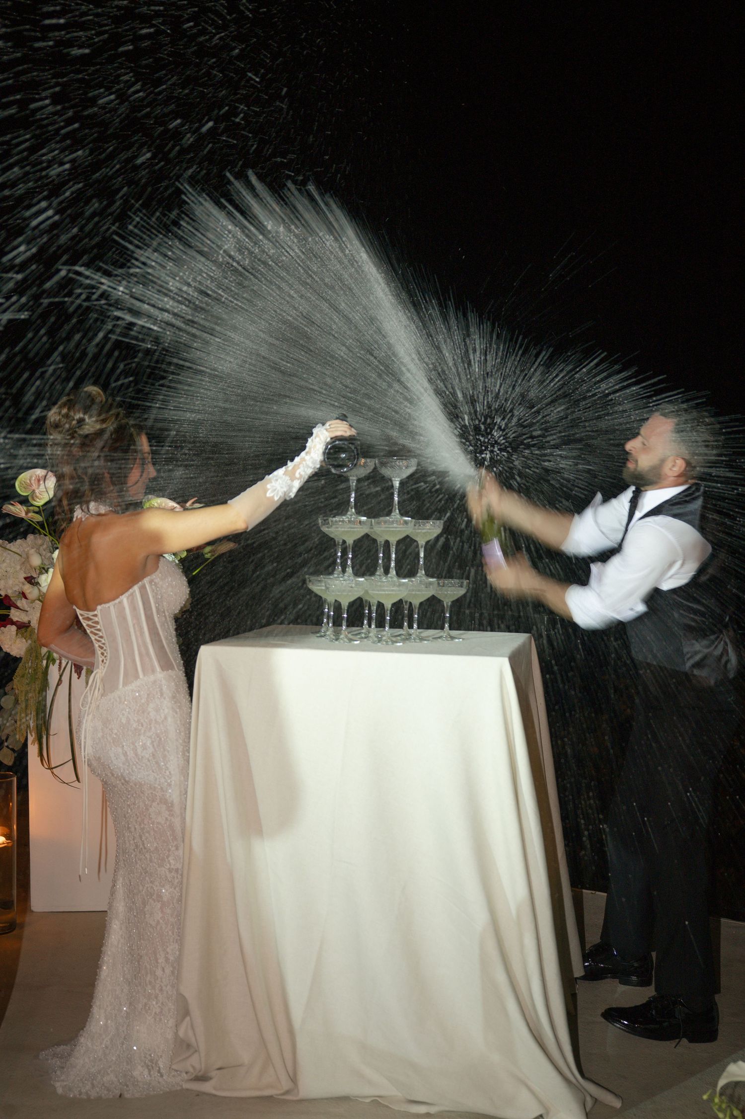 Bride and groom spraying champagne tower at outdoor wedding reception at night.