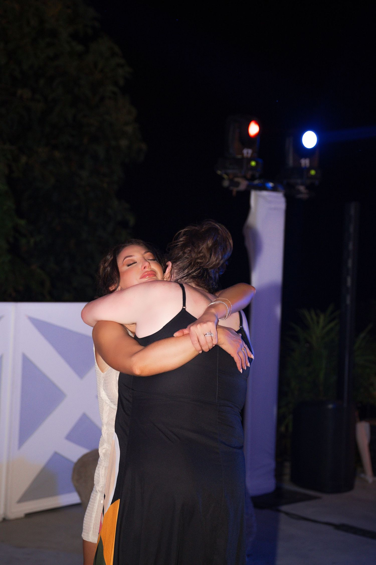 Two women sharing emotional hug on outdoor dance floor at nighttime wedding reception.