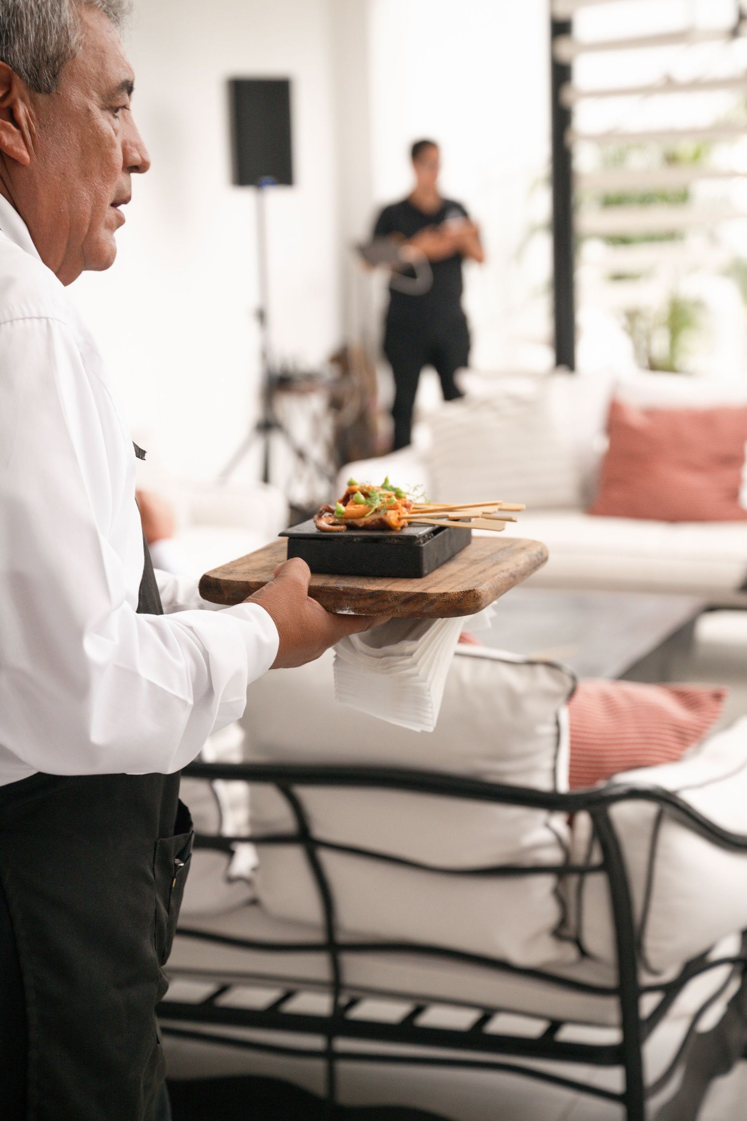 A waiter in white shirt carries a wooden board with gourmet food at an outdoor catered event.