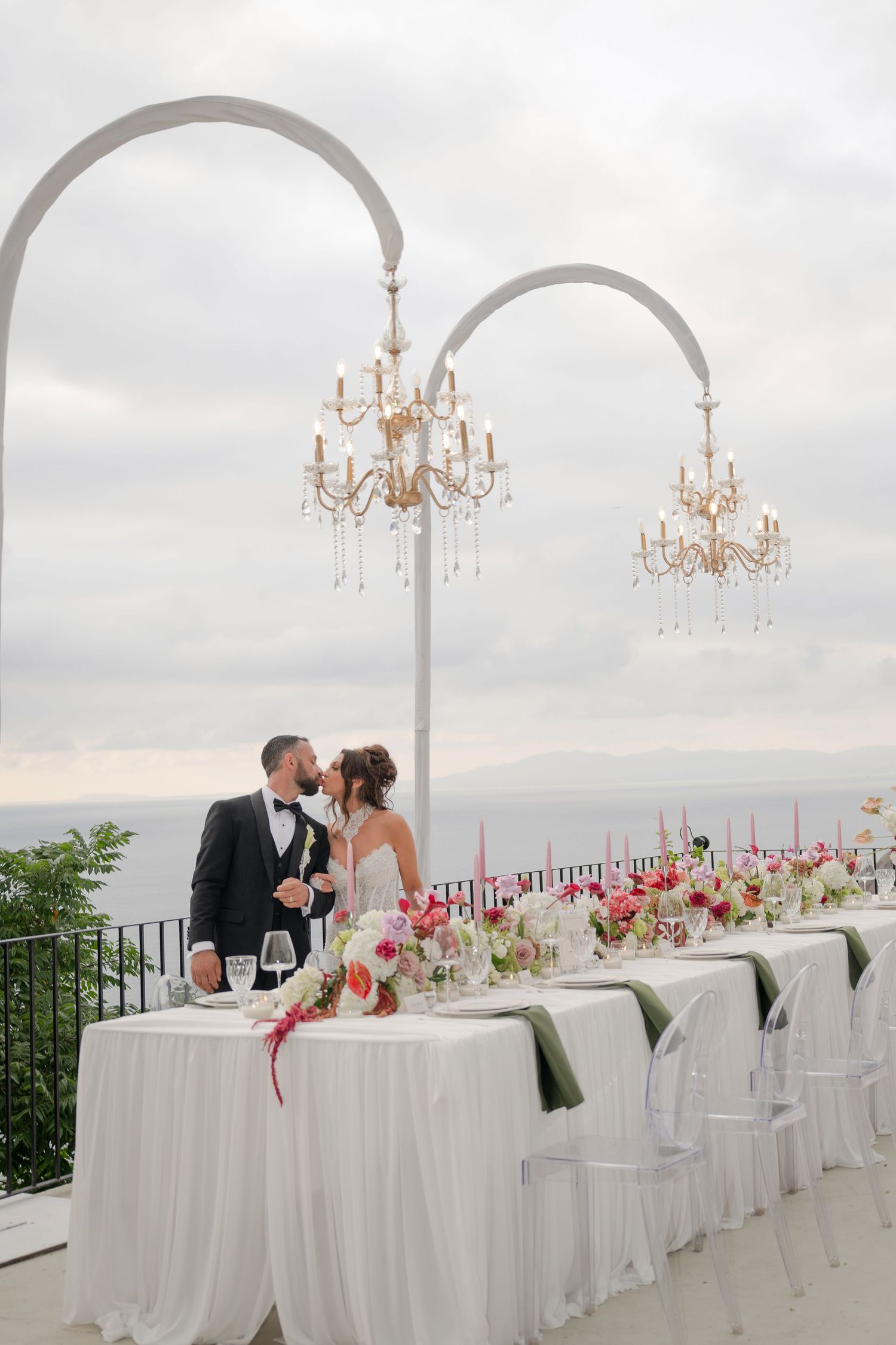 Bride and groom kissing at outdoor oceanview wedding reception table under white arch chandeliers.