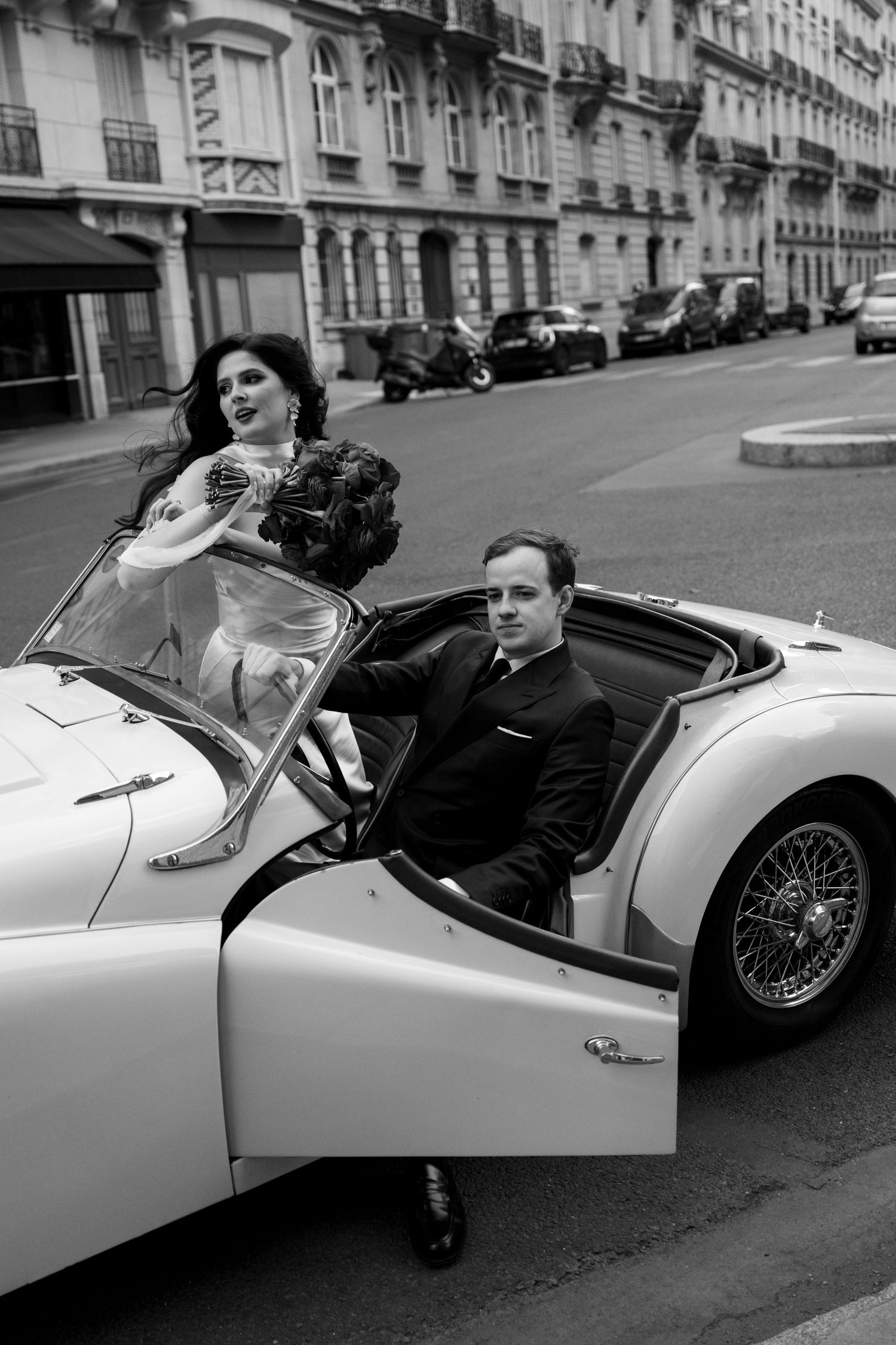 Black and white photo of bride and groom riding in classic white convertible through elegant Paris boulevard.