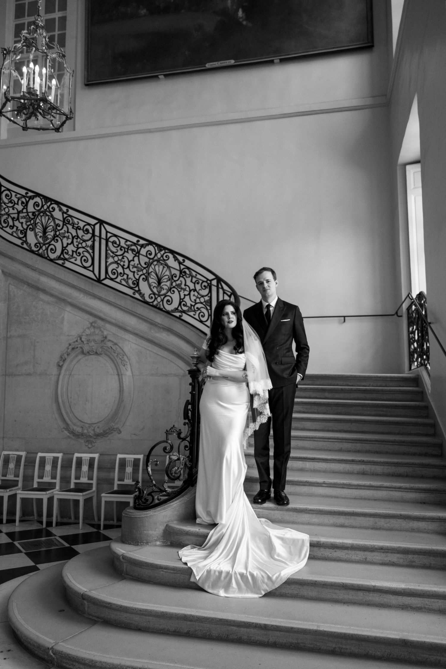 Black and white photo of bride and groom posing on grand ornate staircase with wrought iron railing.