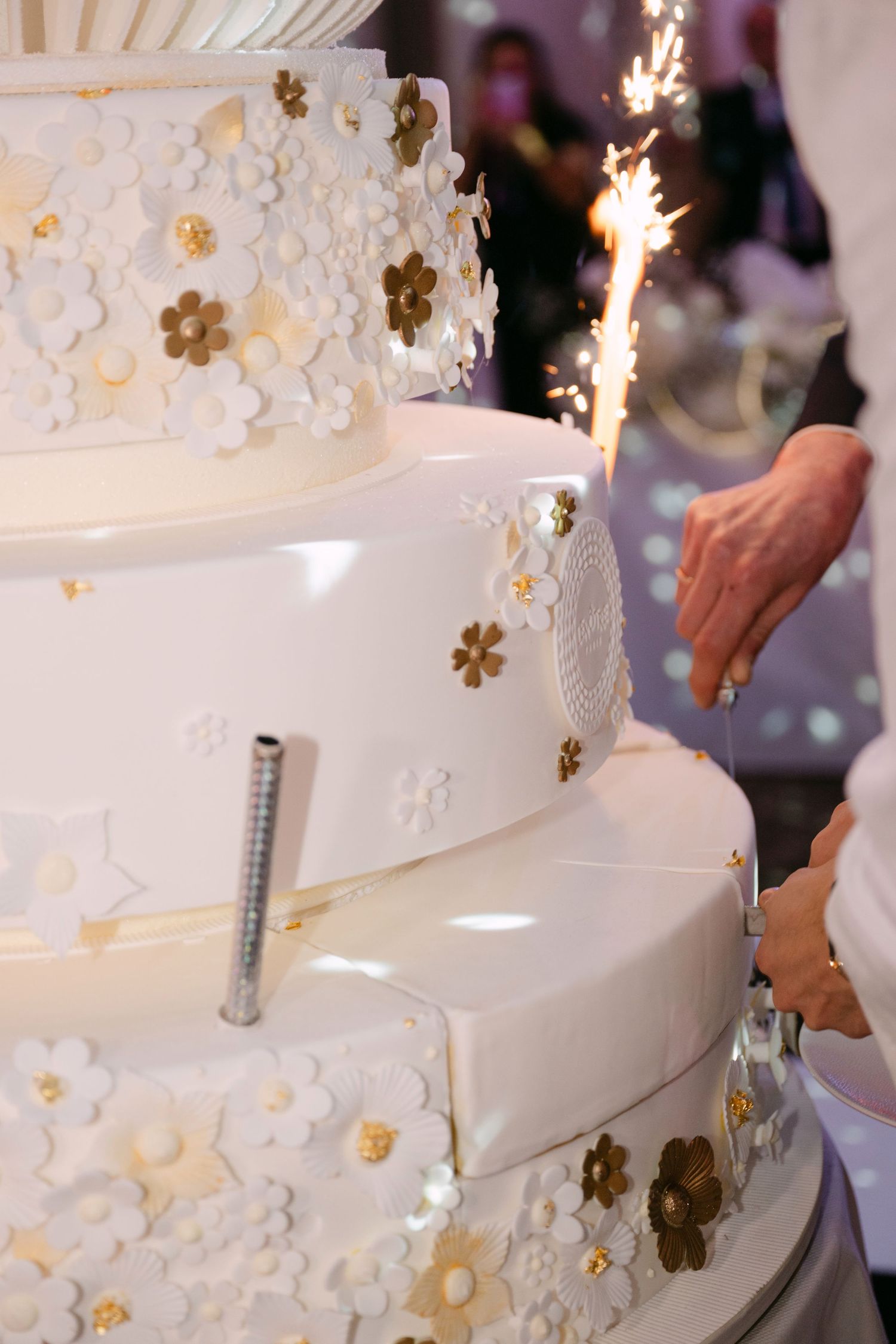 Elegant multi-tier white wedding cake with gold flowers and sparklers being cut at reception.