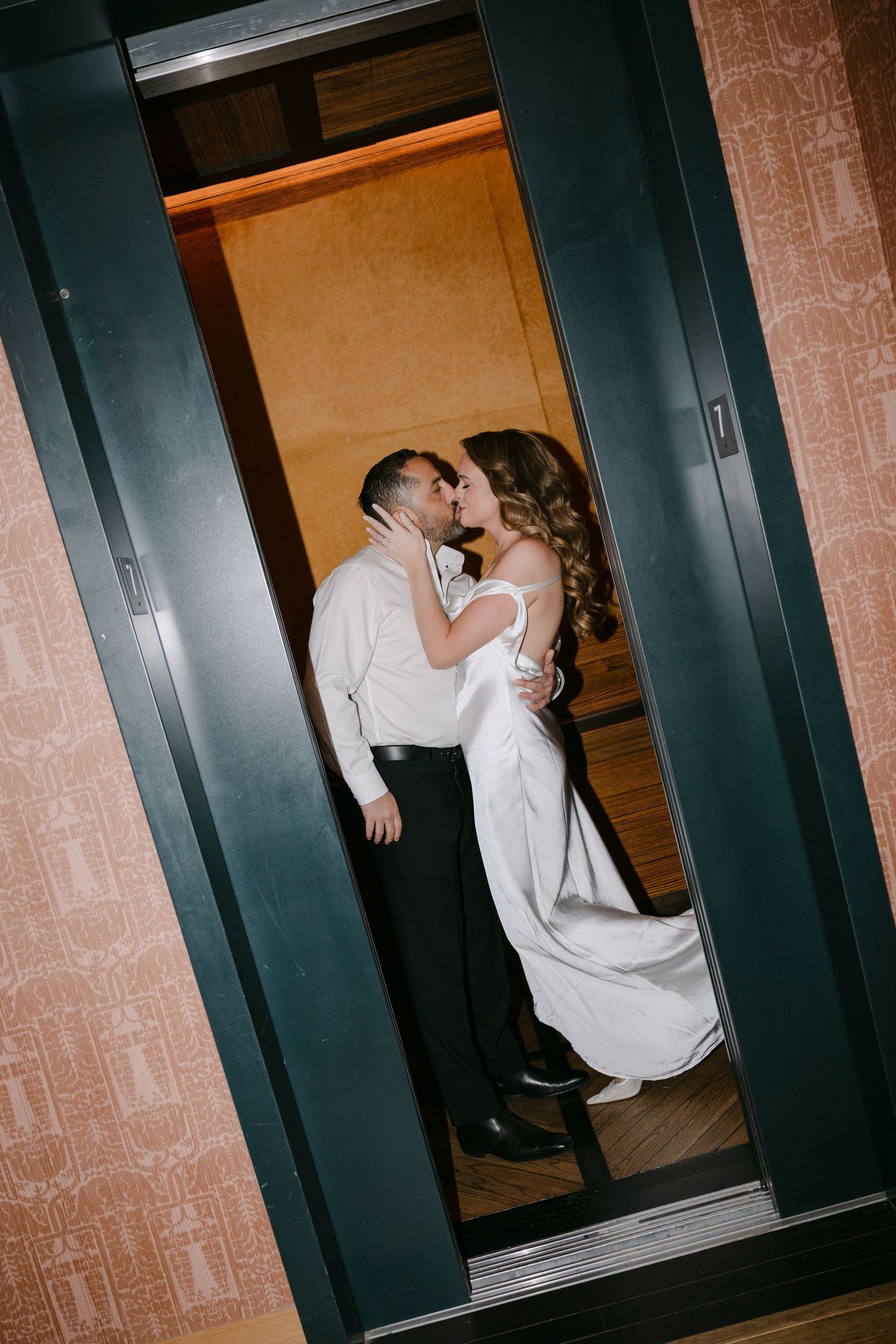 Bride and groom share romantic kiss inside elevator, she wears elegant white satin wedding gown.