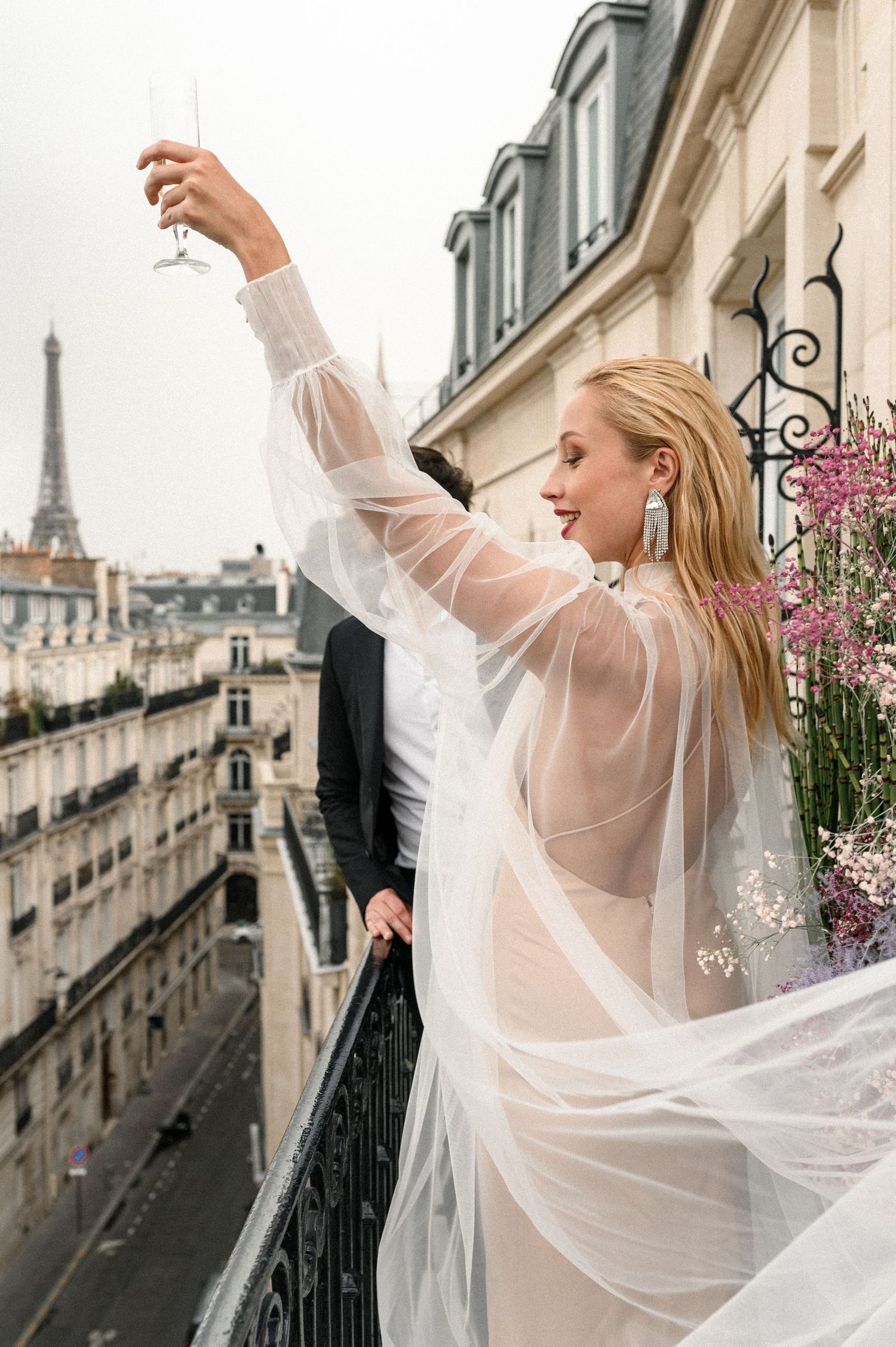 Bride in sheer white gown raising champagne glass on a Paris balcony with Eiffel Tower view.