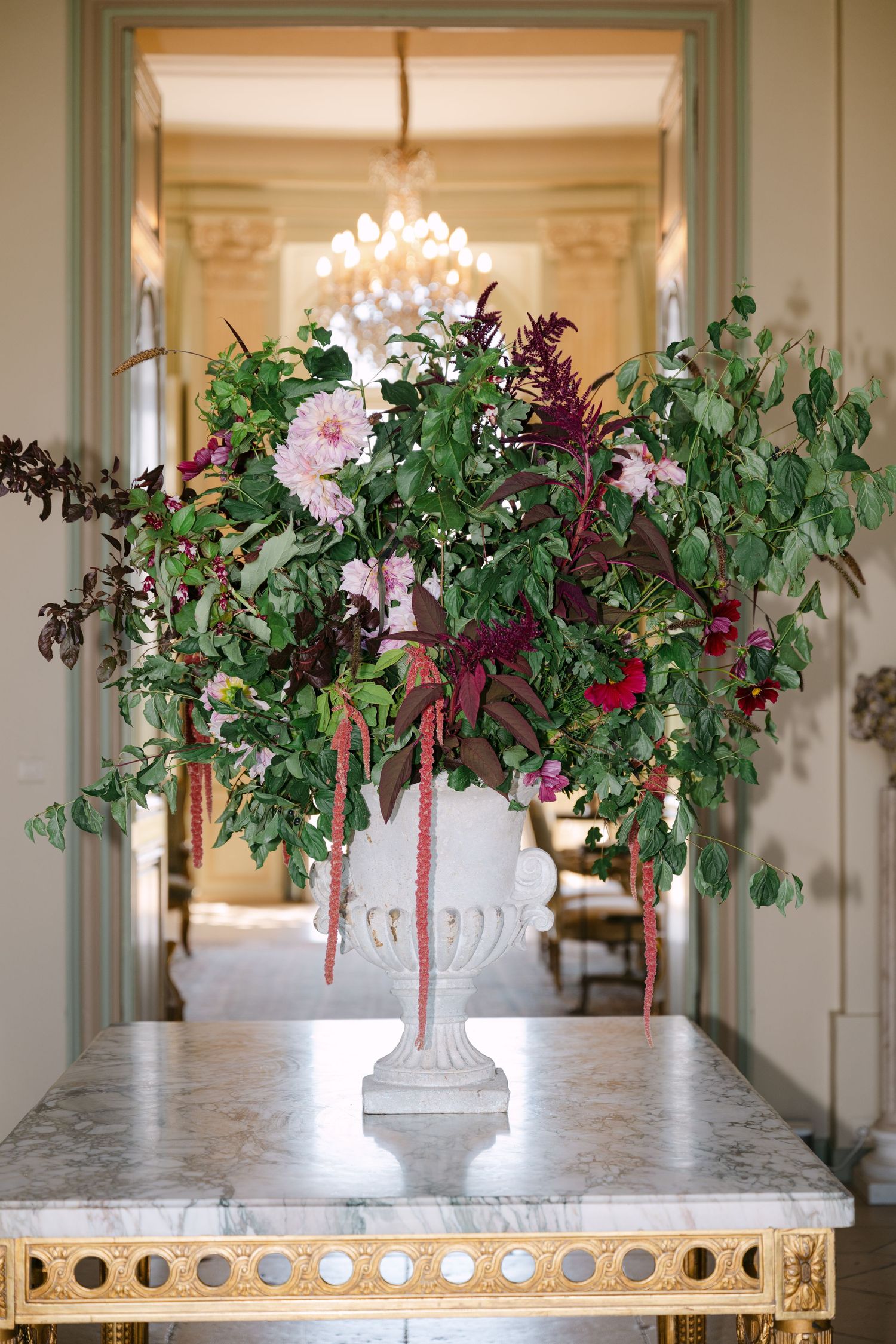 Lush floral arrangement of greenery, dahlias, and burgundy blooms in a white urn on a marble console table.