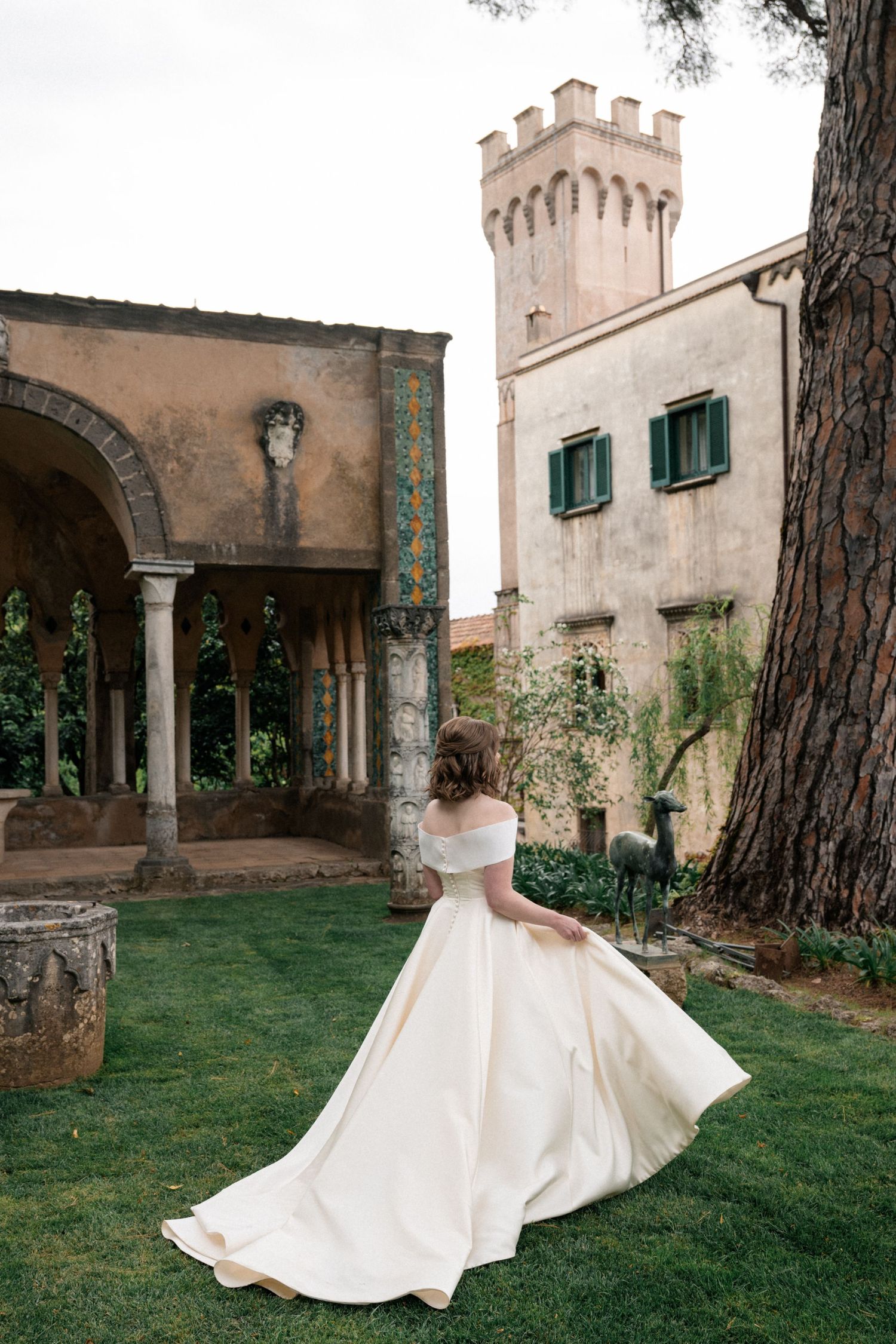 Bride in off-shoulder ball gown twirling in garden of historic Italian villa with tower.