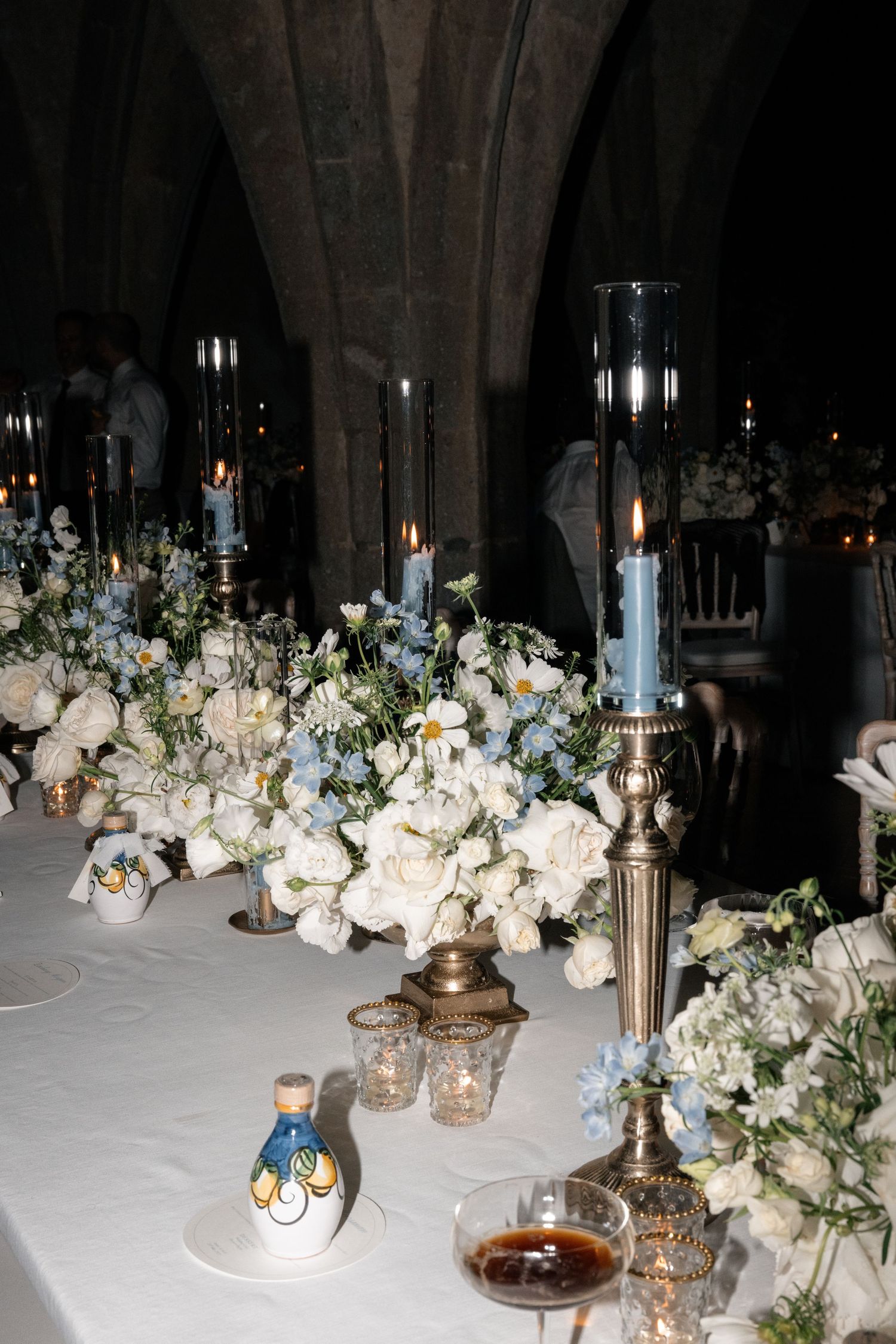 Elegant wedding reception table adorned with white roses, blue candles, and gold candelabras in a stone venue.