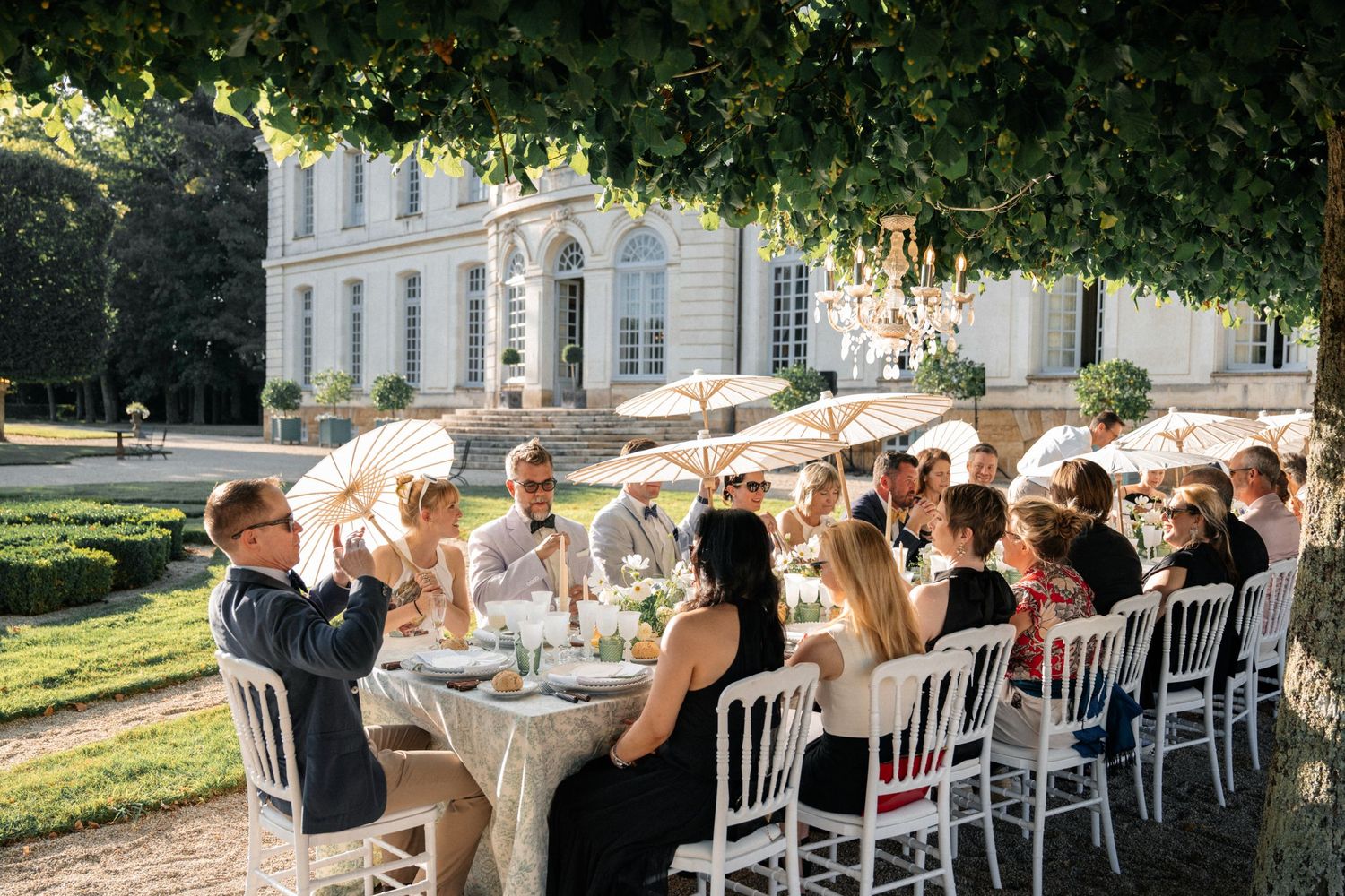 Wedding guests dining at an outdoor reception table beneath trees in front of a classic French chateau.