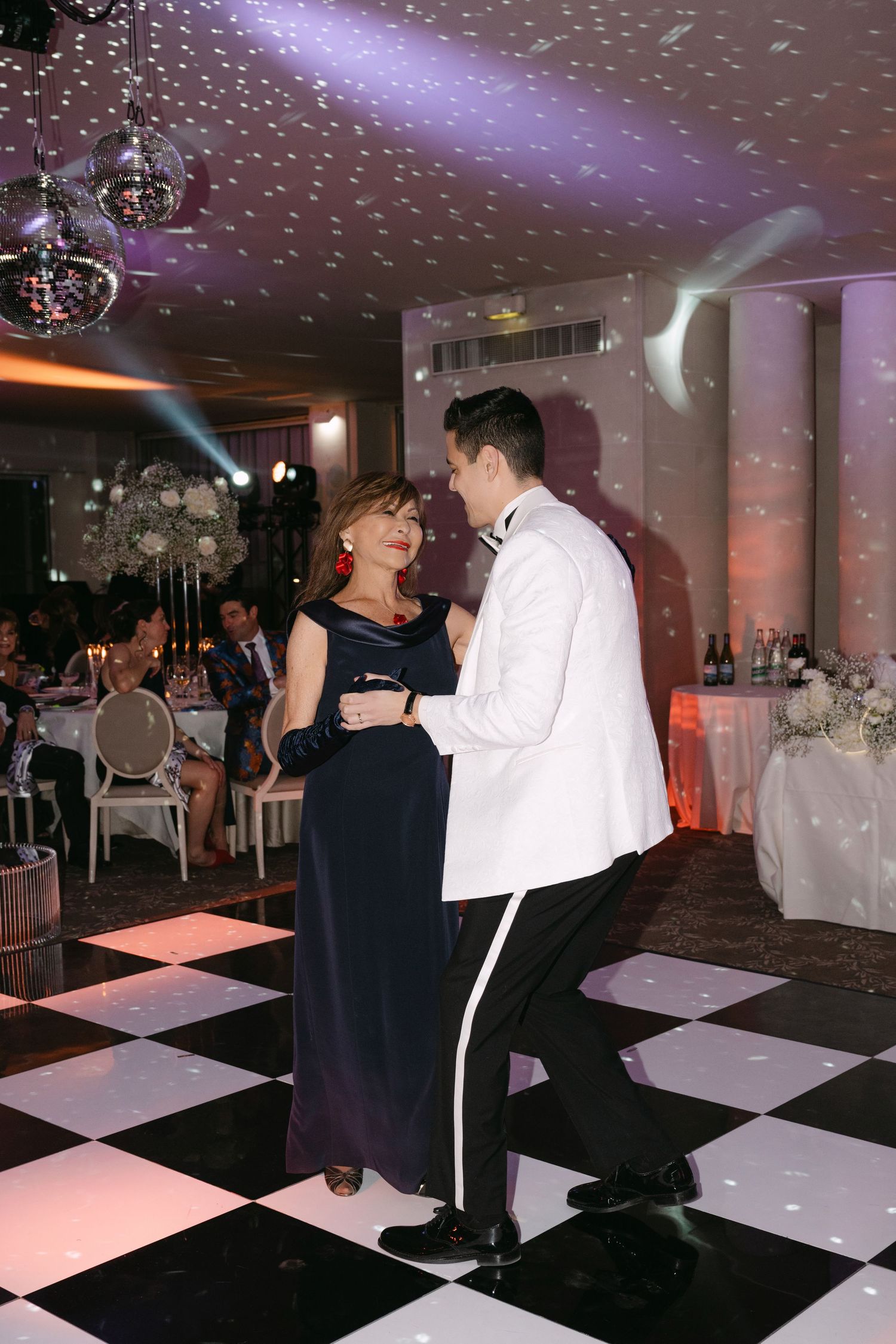Groom in white tuxedo shares mother-son dance on checkered floor at elegant wedding reception.