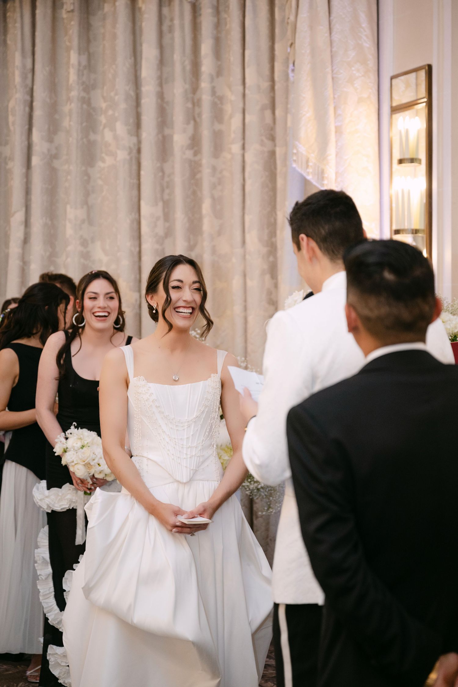 Bride laughing joyfully at altar during wedding ceremony while bridesmaid smiles behind her.