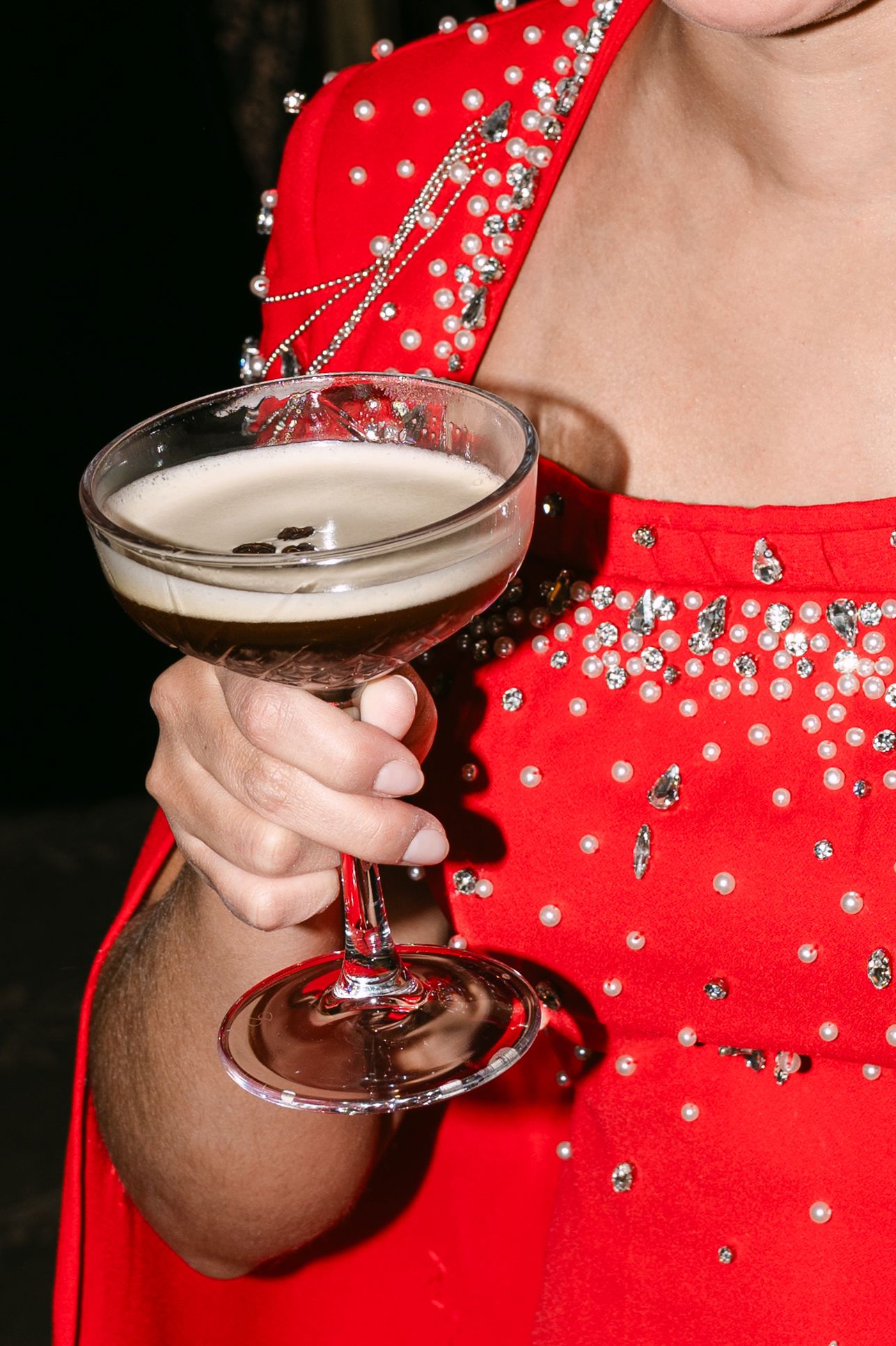 Close-up of guest in red beaded gown holding espresso martini cocktail at formal wedding reception event.