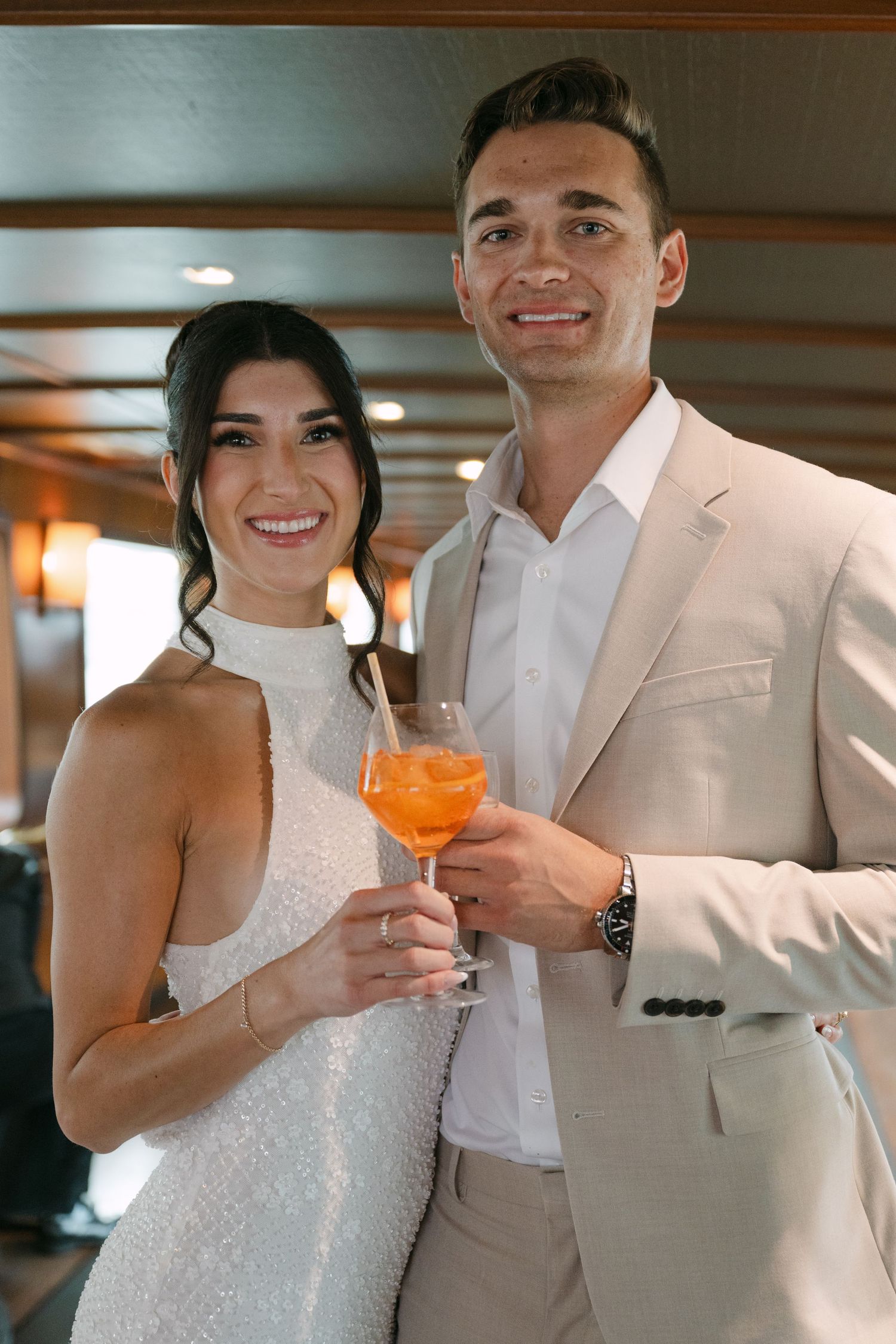 A smiling bride in white and groom in beige suit share an Aperol Spritz inside a luxury boat.