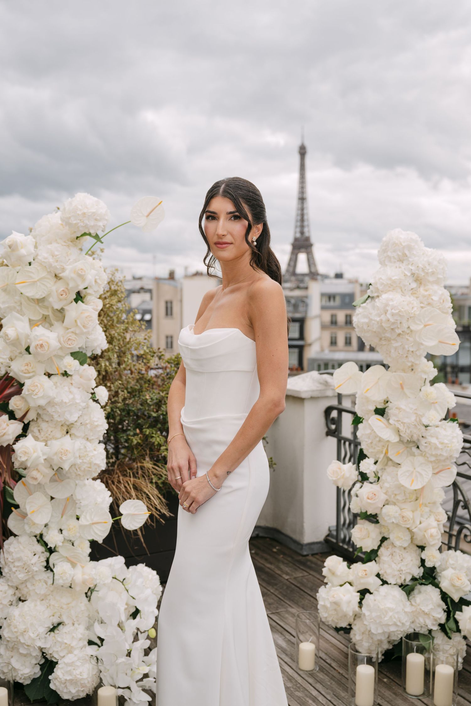 Elegant bride in strapless white gown poses between white floral columns with Eiffel Tower in background.