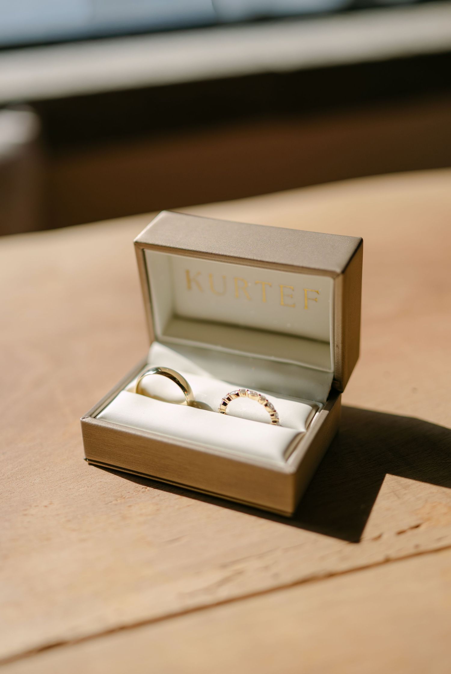 Two gold wedding bands in a Kurtér jewelry box, one plain and one with gemstones, resting on a wooden surface.