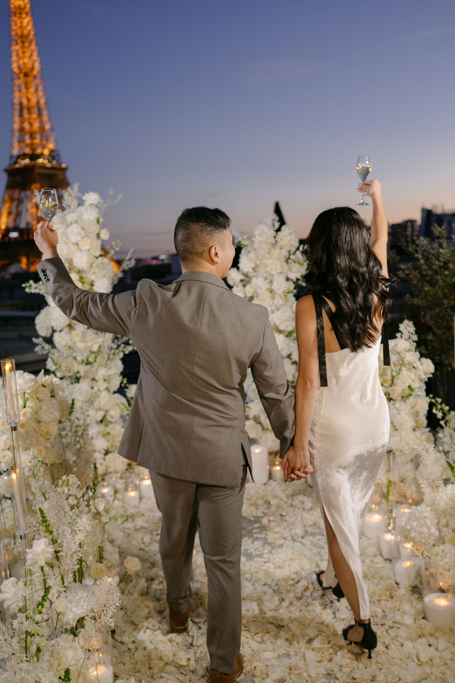 Couple raising champagne glasses amid white flowers and candles at dusk with the glowing Eiffel Tower in Paris behind them.