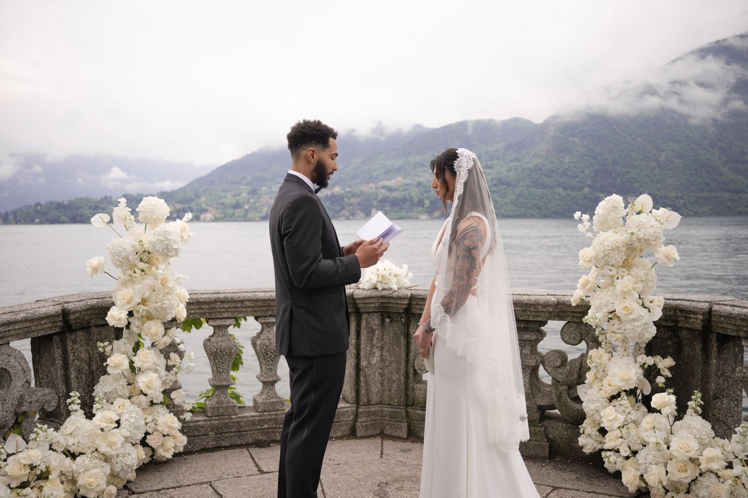 Couple exchanging vows on a lakeside terrace adorned with white floral arrangements, mountains in the background.