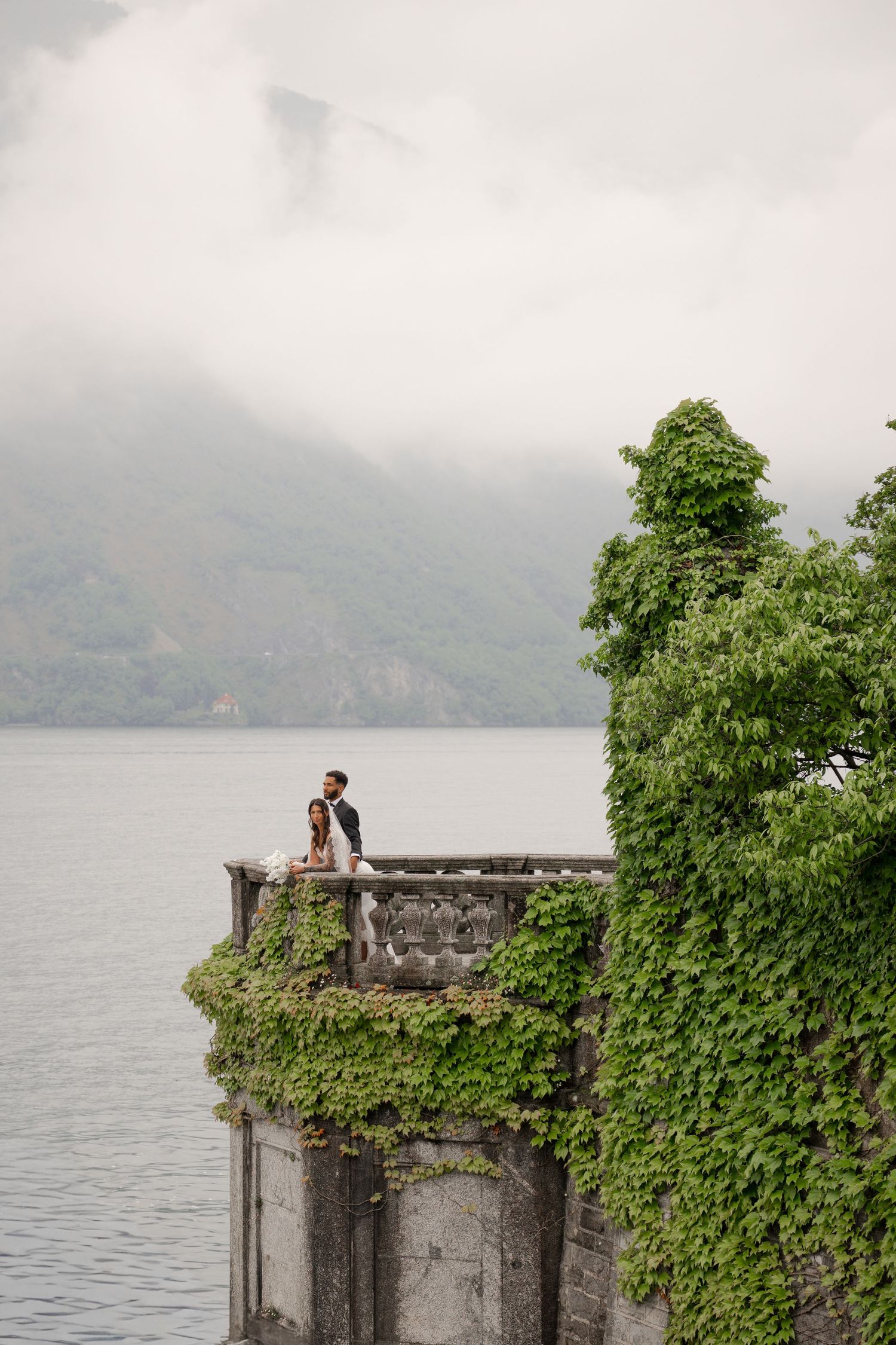 Newlyweds standing on ivy-covered stone balcony overlooking misty lake and mountain landscape.