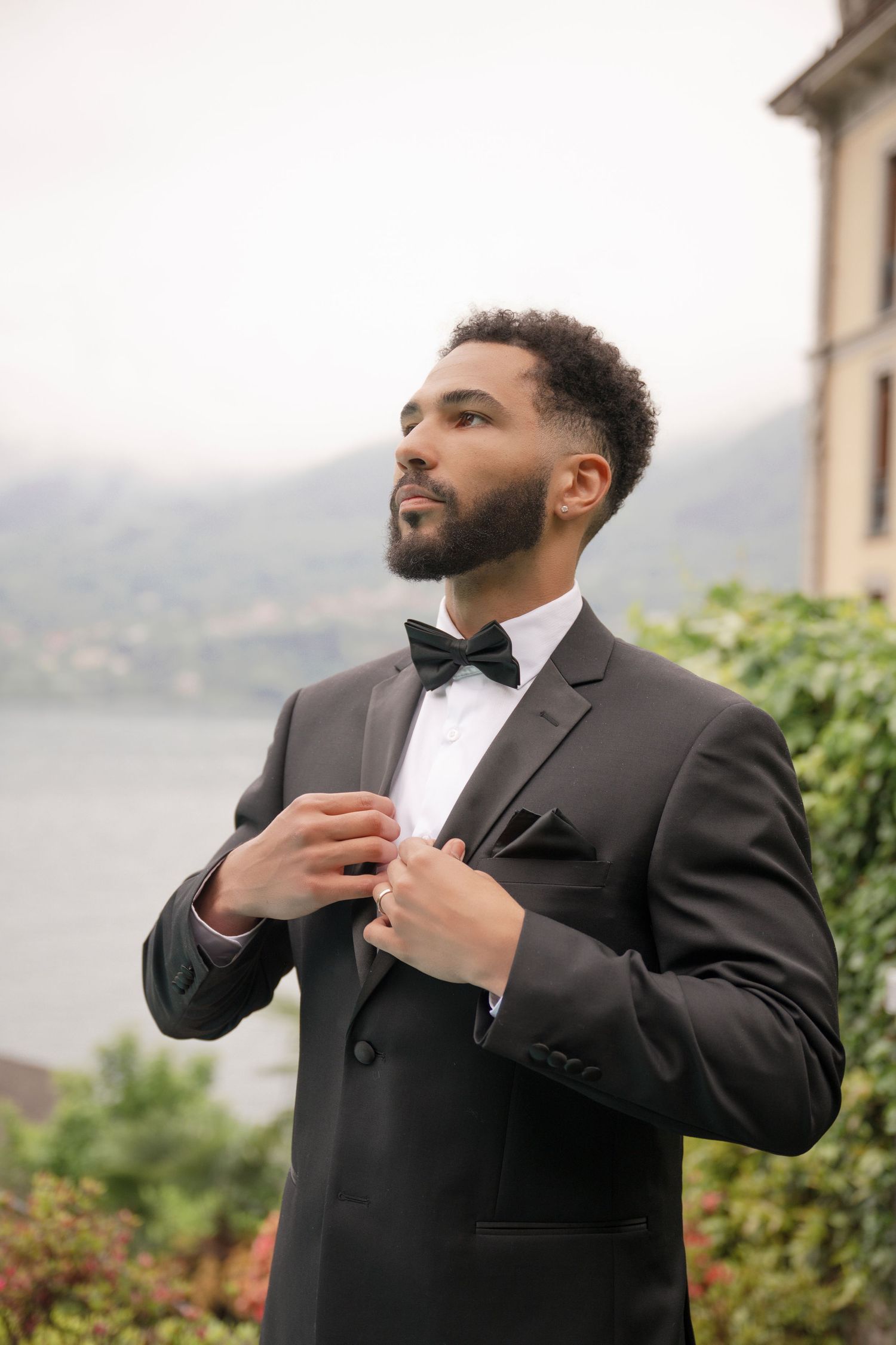 Handsome groom in black tuxedo adjusting jacket with scenic lake and mountains in background.