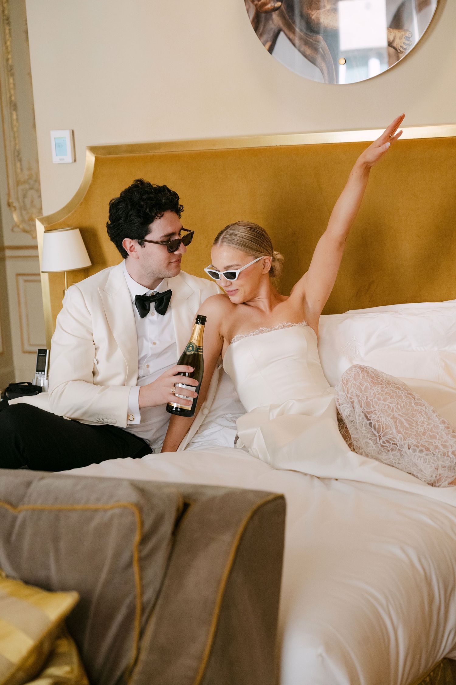 Newlyweds in sunglasses celebrate on hotel bed with champagne, bride in strapless gown raising arm joyfully.