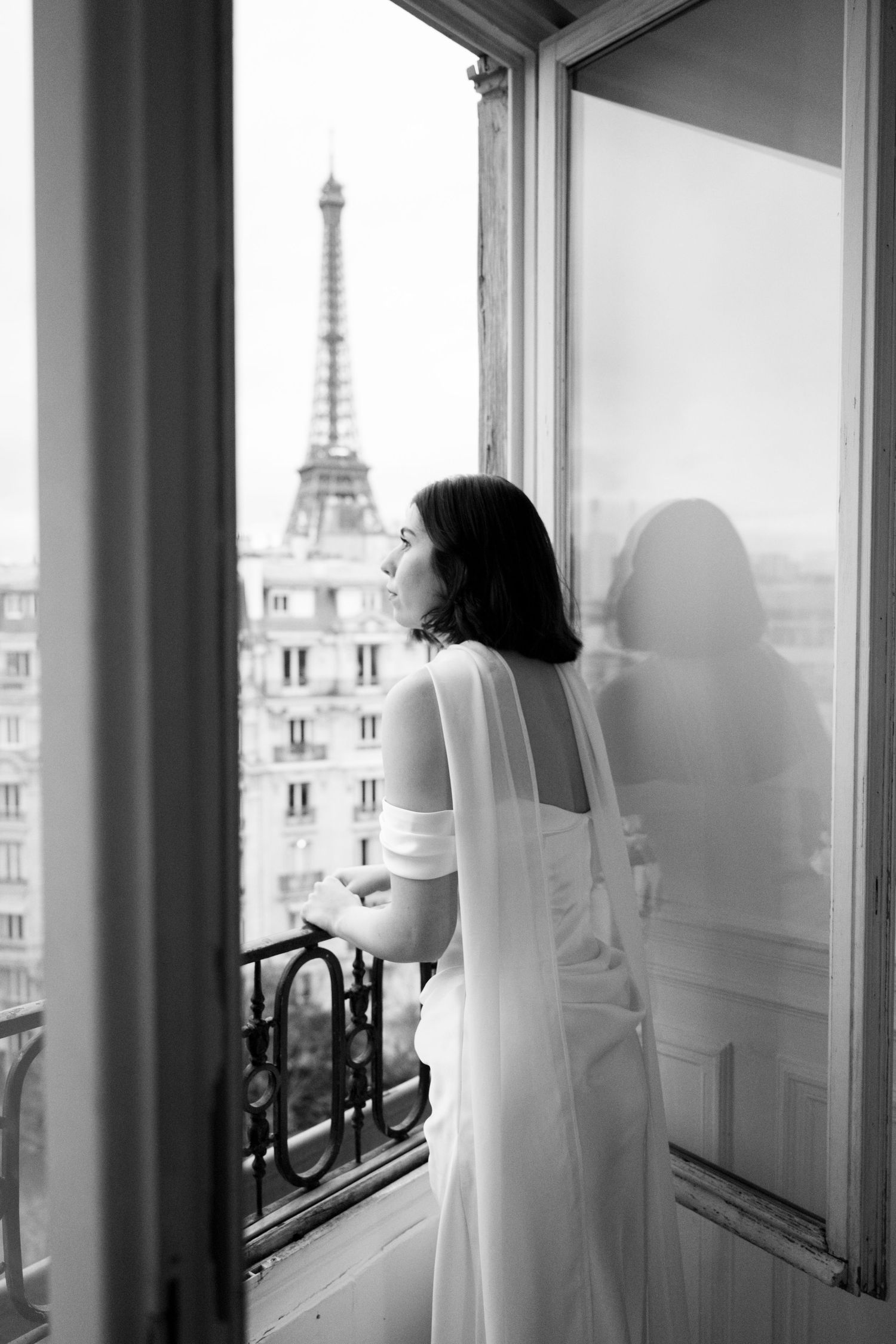 Black and white photo of a bride in white dress gazing at the Eiffel Tower from a Parisian balcony window.