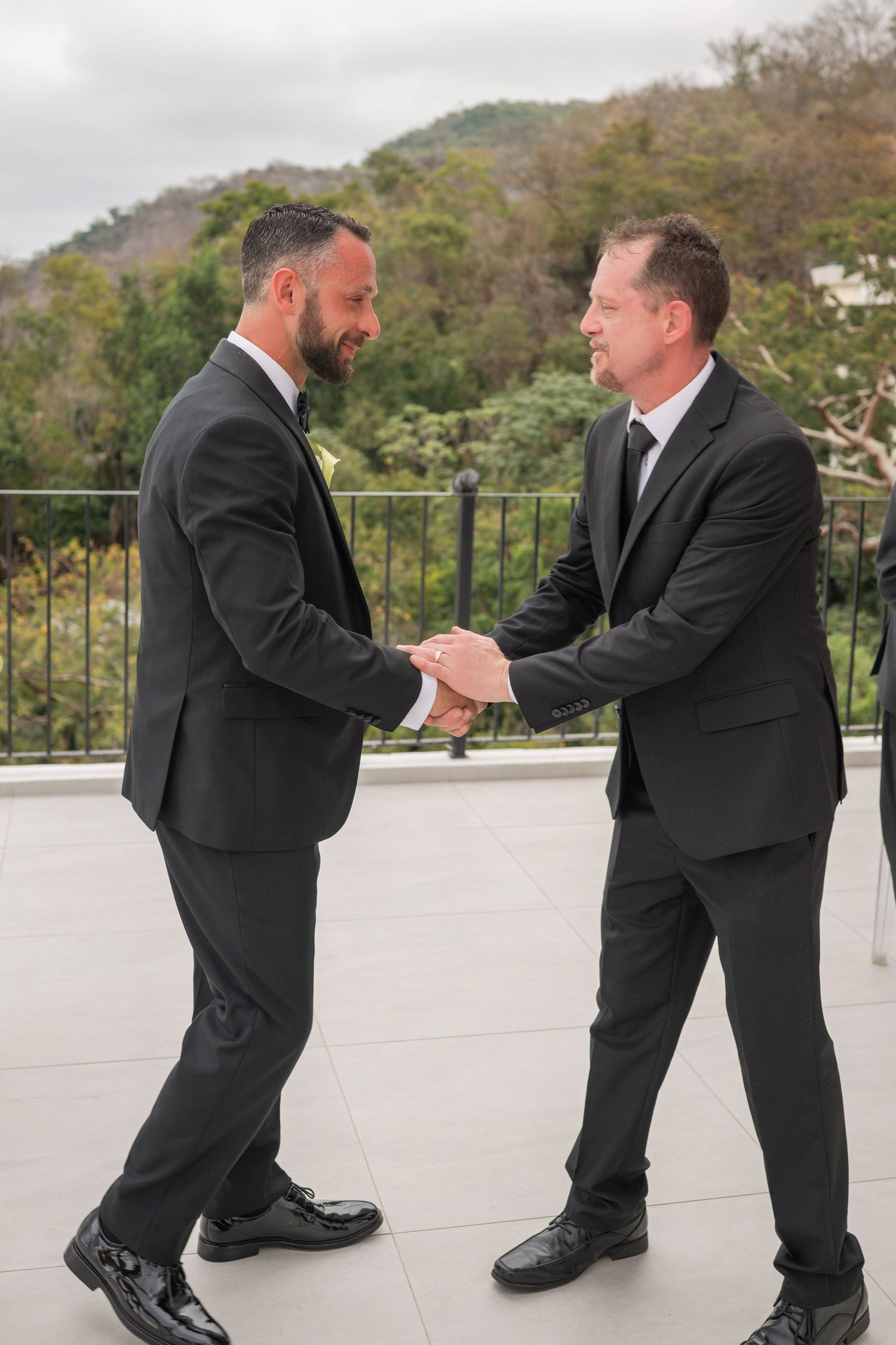 Two men in black suits shaking hands on an outdoor terrace with lush green hills in the background.