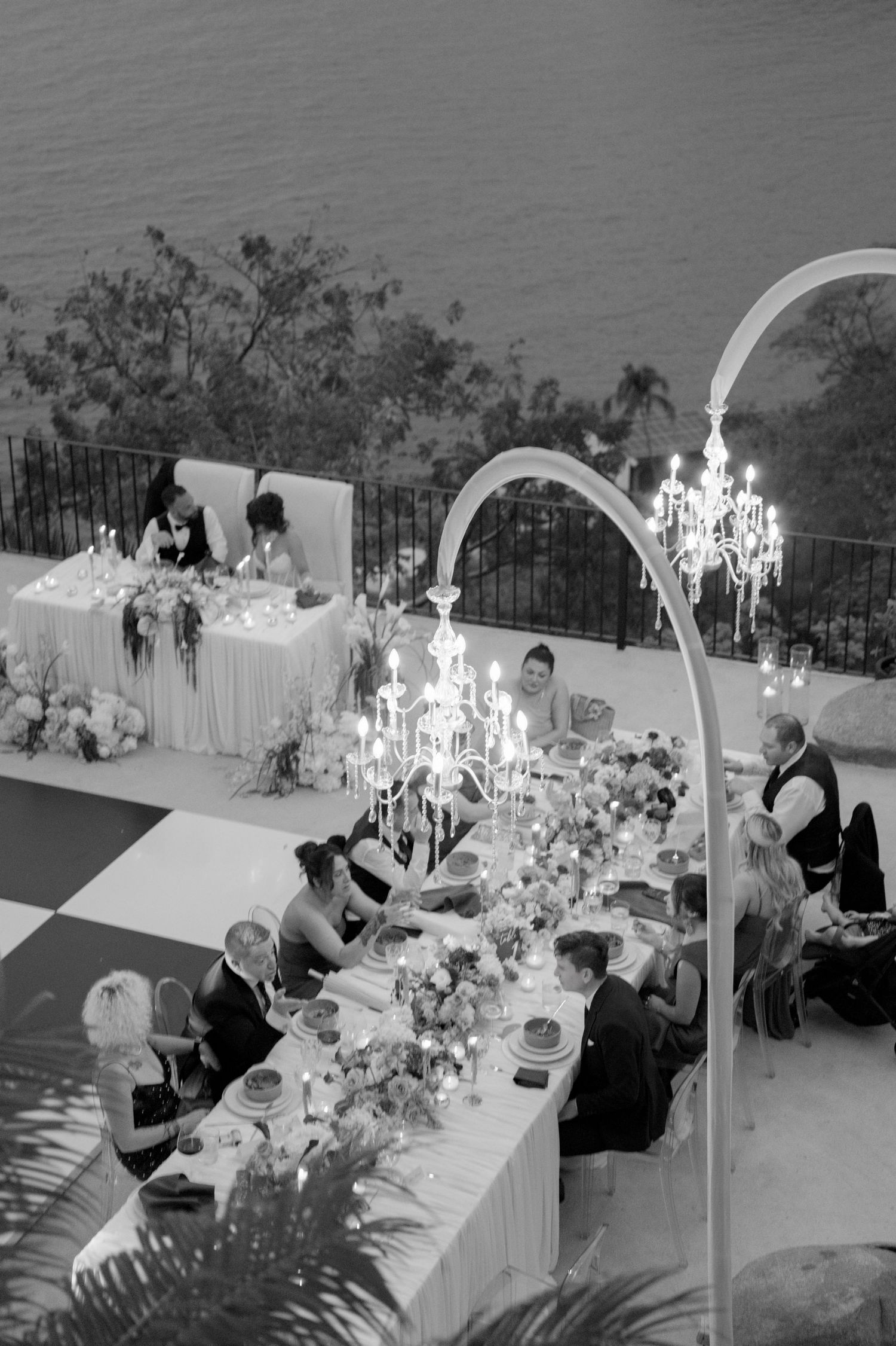 Aerial black and white view of elegant outdoor wedding reception with chandelier and ocean backdrop.