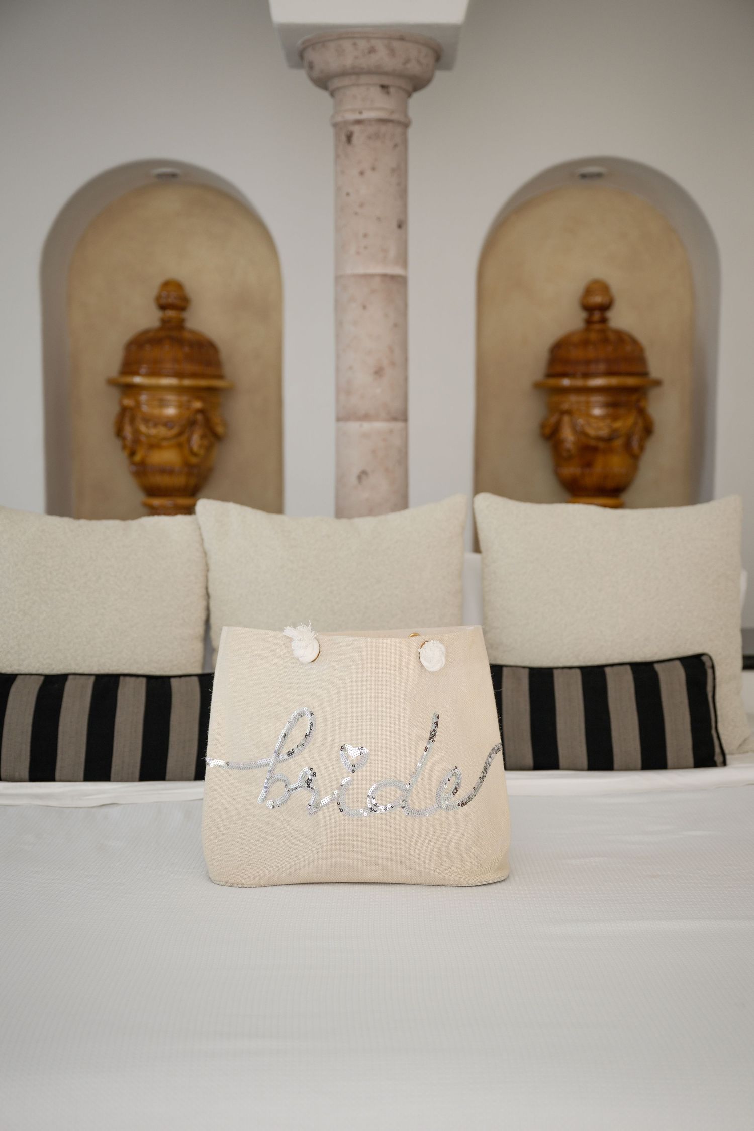 A decorative bride pillow with silver sequin lettering on a white bed with striped pillows and ornate golden urns in wall niches.