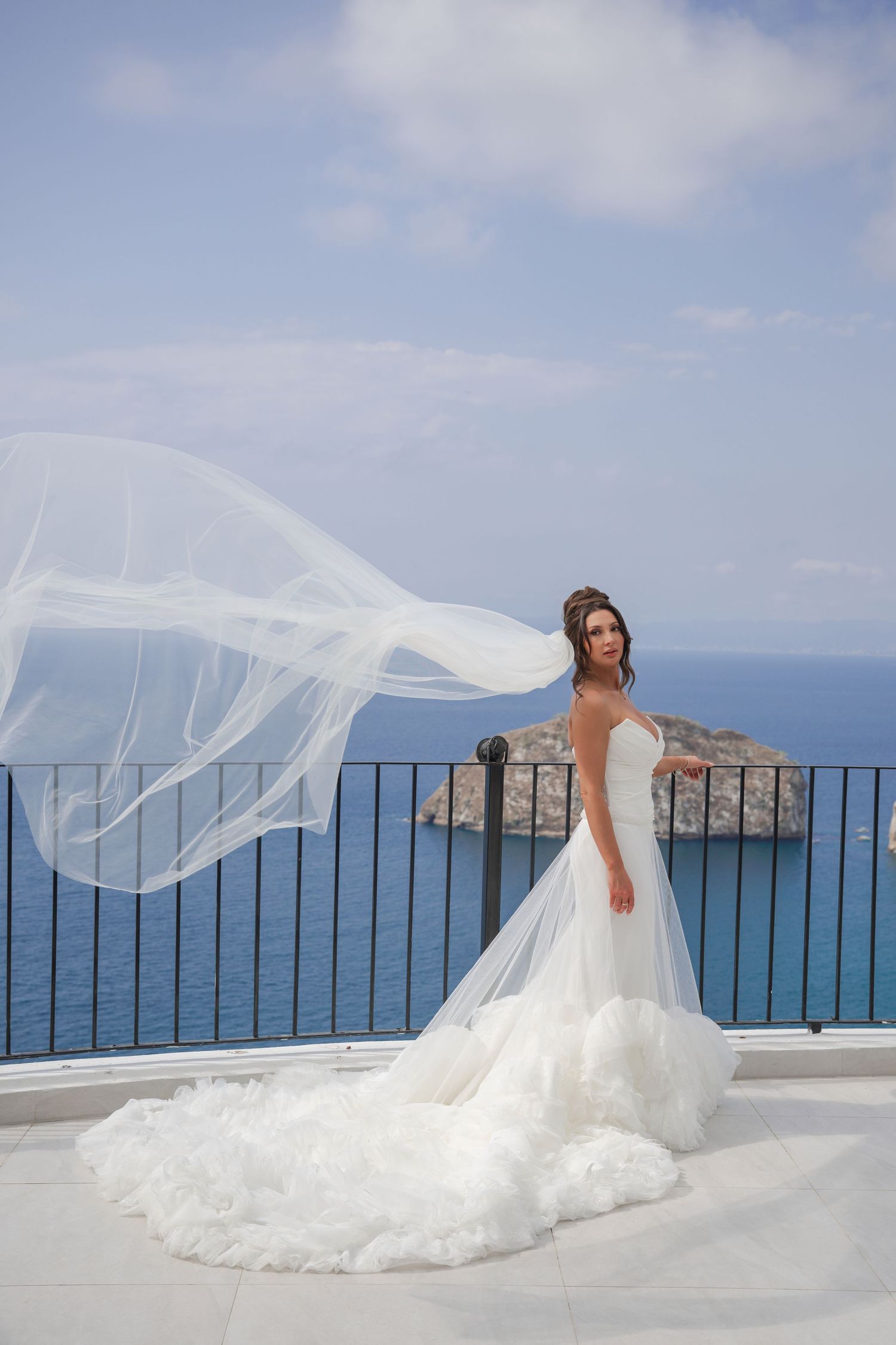 Bride in white wedding gown with flowing veil billowing in wind on seaside terrace.