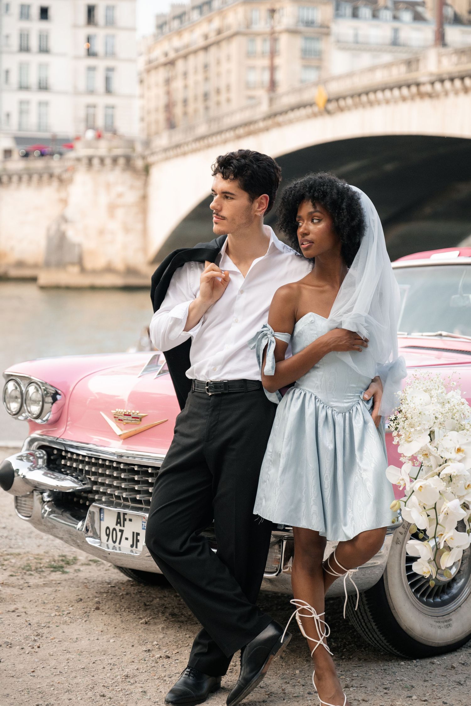 Stylish interracial couple lean against a vintage pink Cadillac beside the Seine River in Paris.