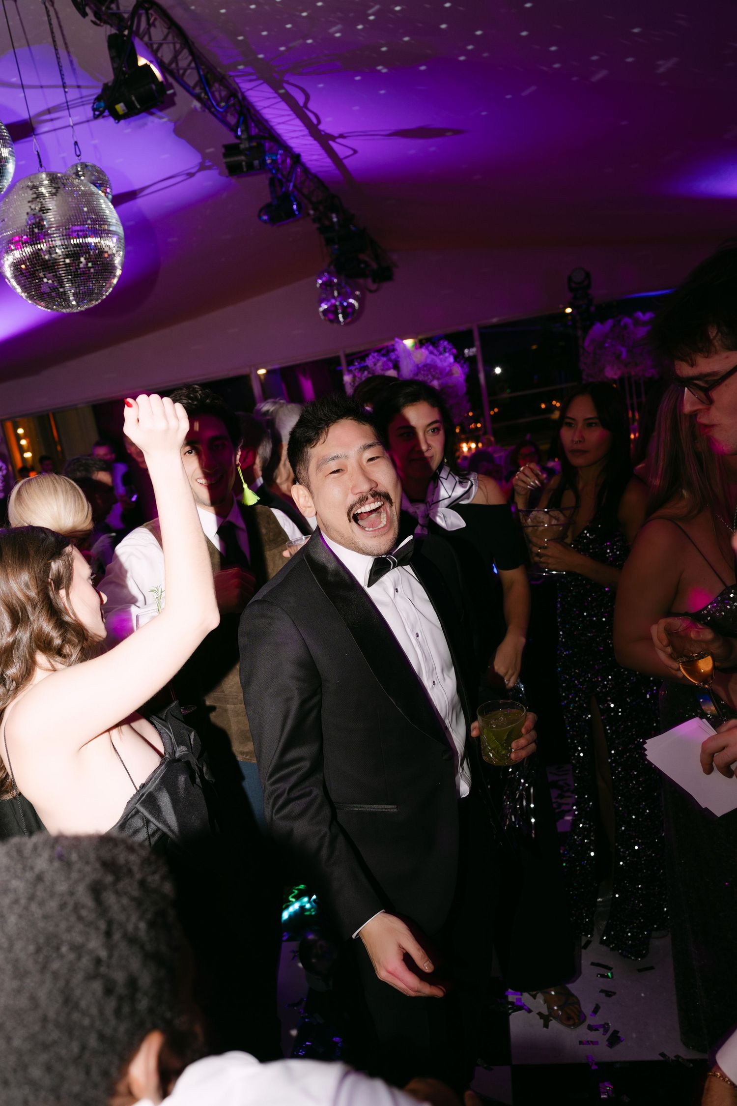 Man in black tuxedo laughing and celebrating on dance floor under disco ball at formal wedding reception.