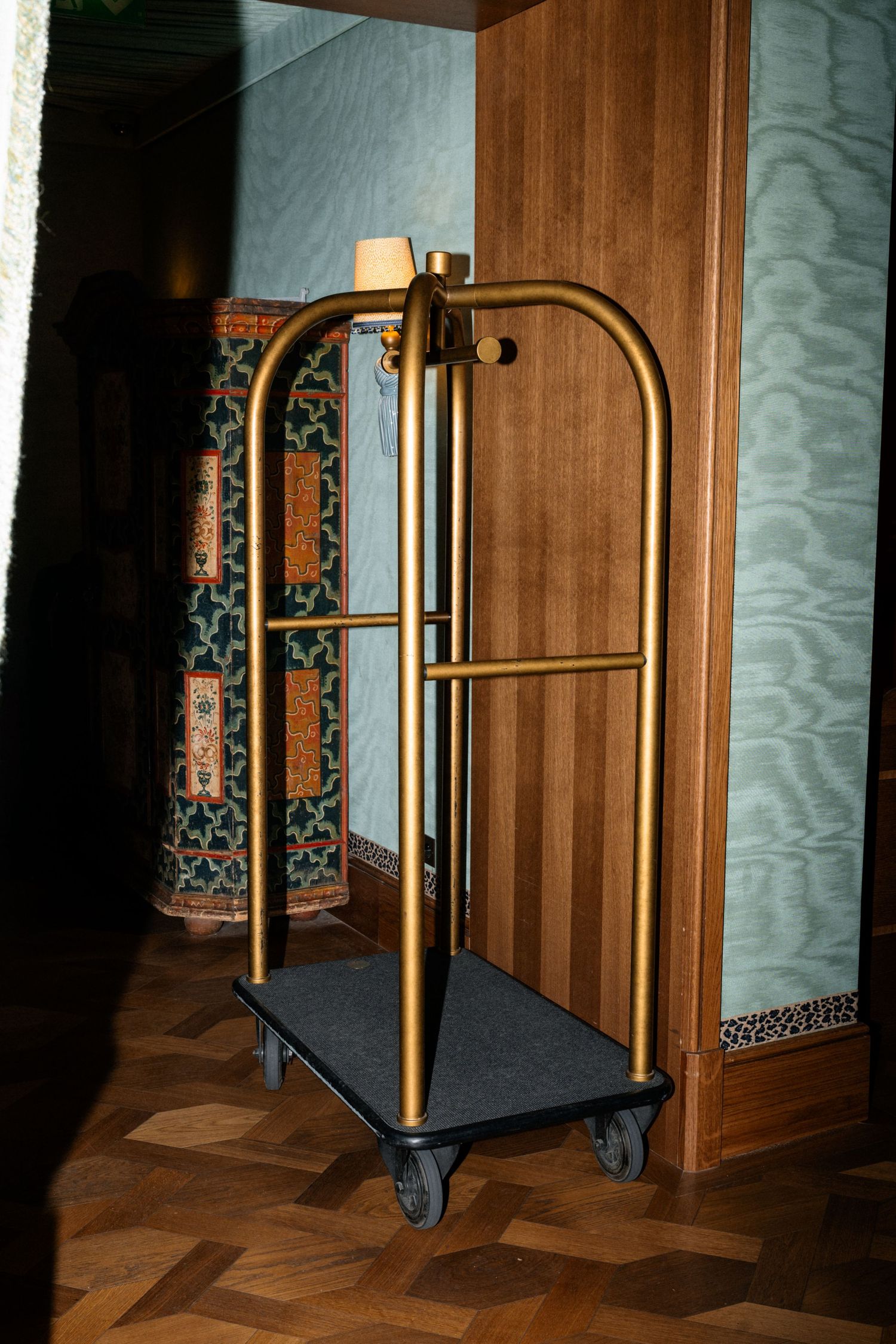 Gold hotel luggage cart in an elegant corridor with patterned wallpaper and wood paneling.