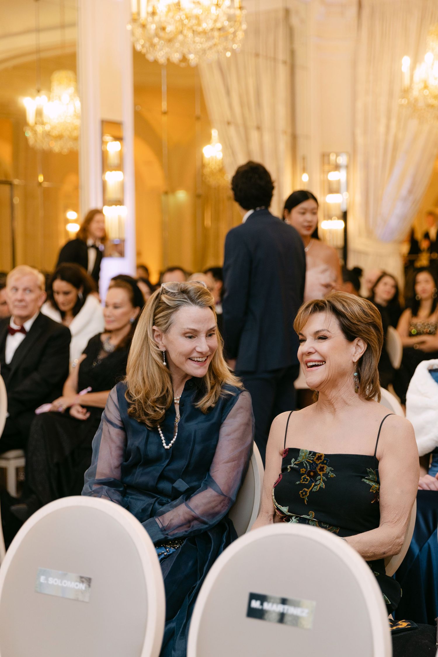 Wedding guests in formal attire seated in an elegant ballroom, two women in navy smiling during the ceremony.
