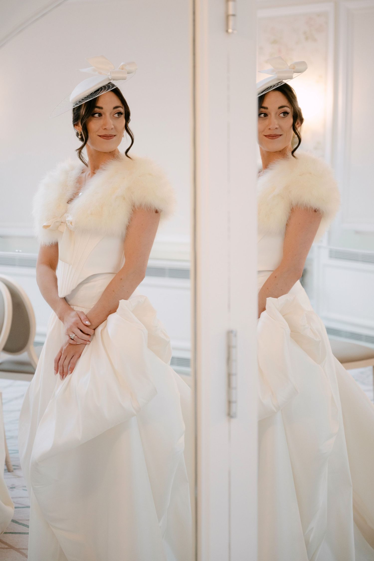 Elegant bride in white ball gown with fur shrug and fascinator hat reflected in full-length mirror.
