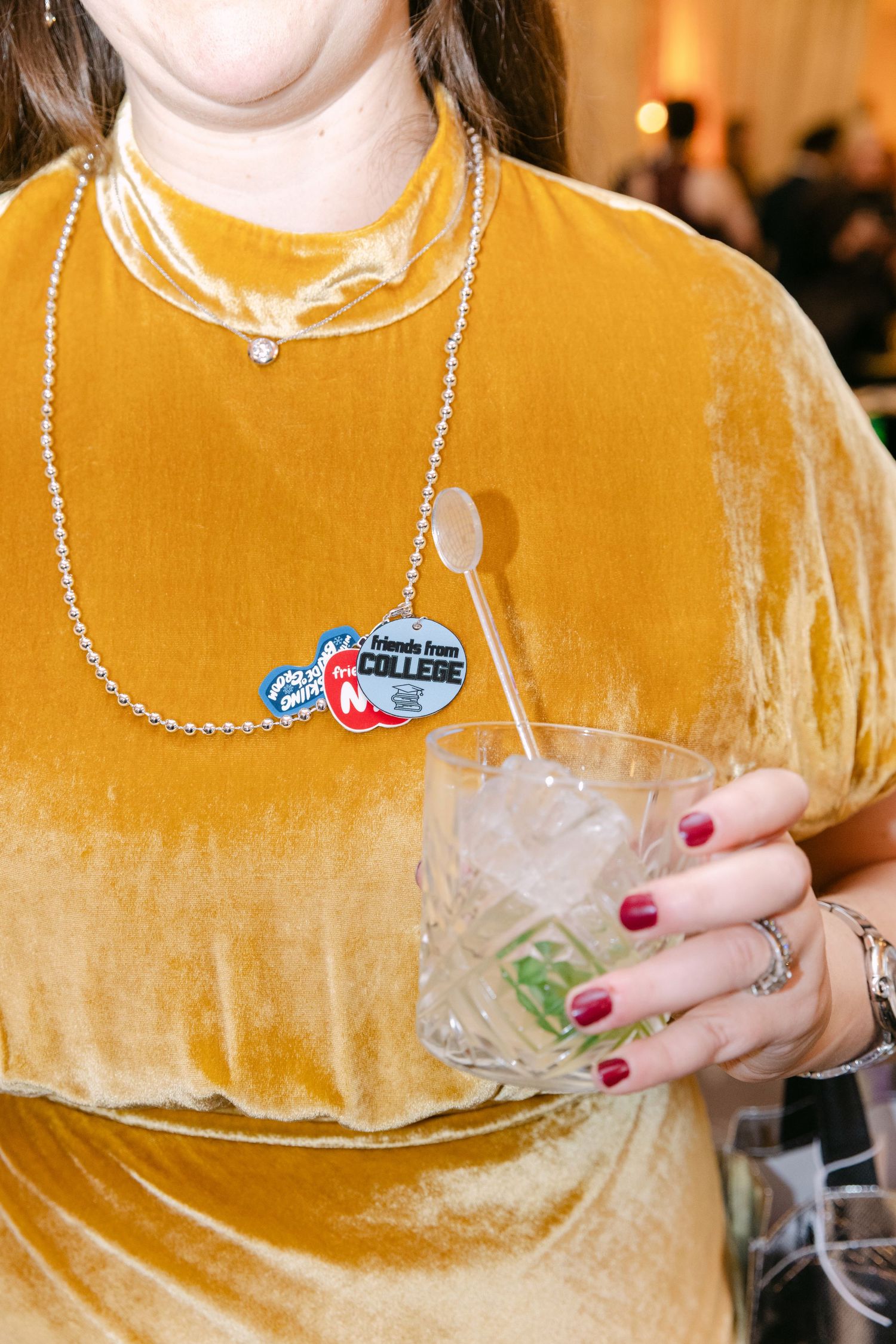 Woman in gold velvet dress holding cocktail glass with silver bead necklace and colorful pins at party.