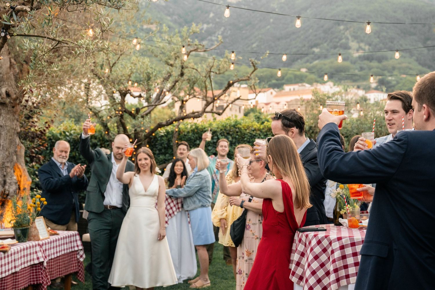 Joyful wedding reception outdoors with bride, groom, and guests celebrating under string lights in garden.