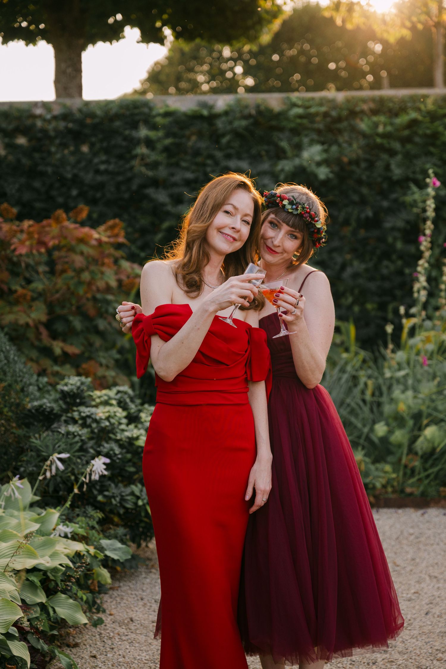 Two women in red and burgundy dresses holding cocktails smiling at sunset garden wedding.