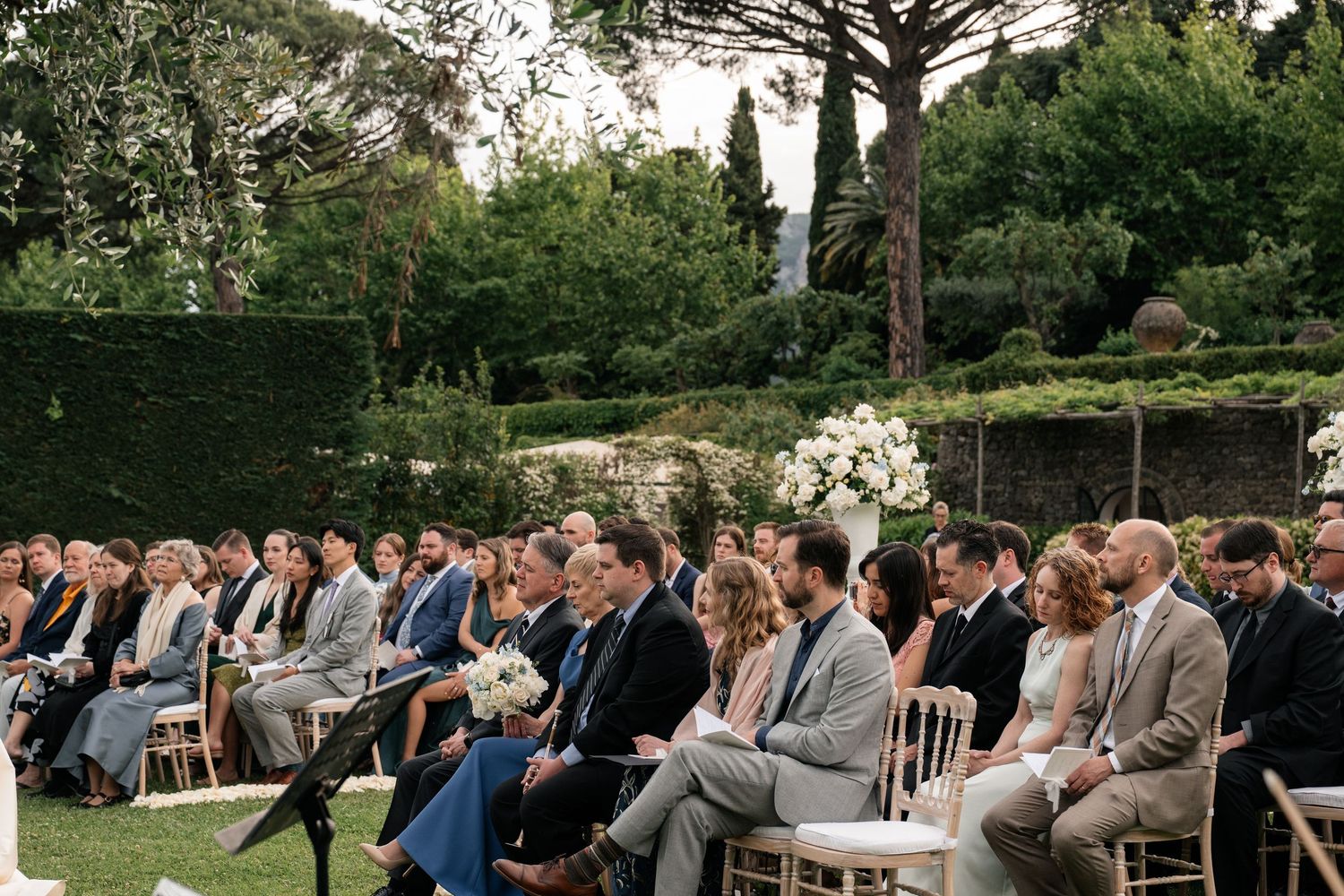 Wedding guests seated in gold chairs watching outdoor ceremony in beautifully landscaped Italian garden.