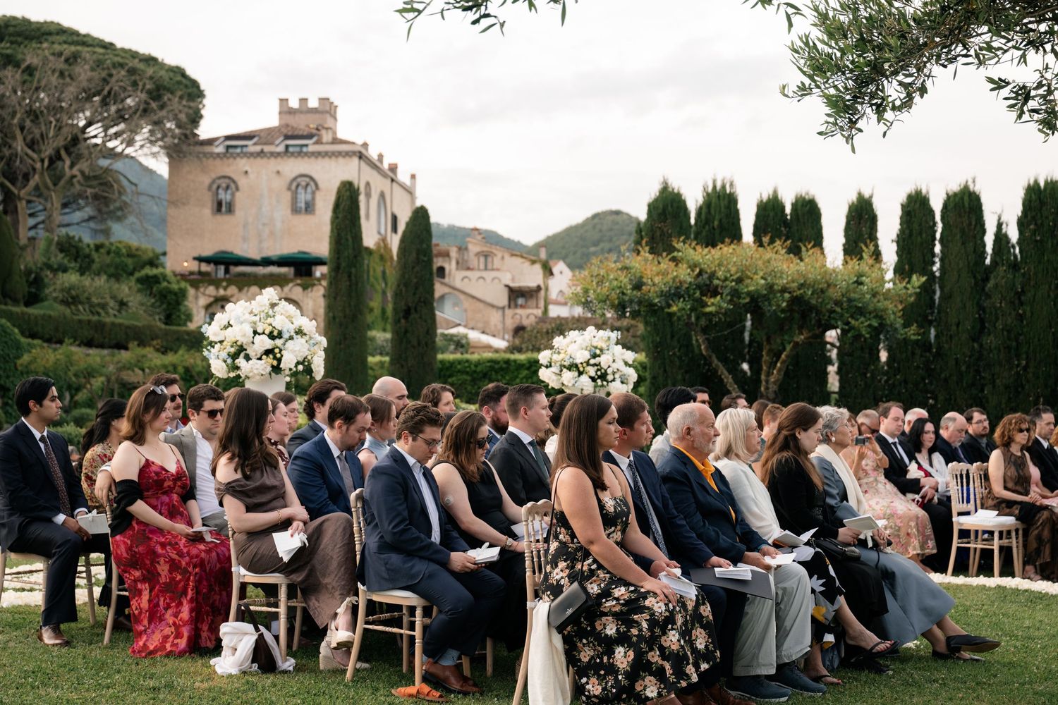 Guests seated at outdoor wedding ceremony with Italian villa and cypress trees backdrop.