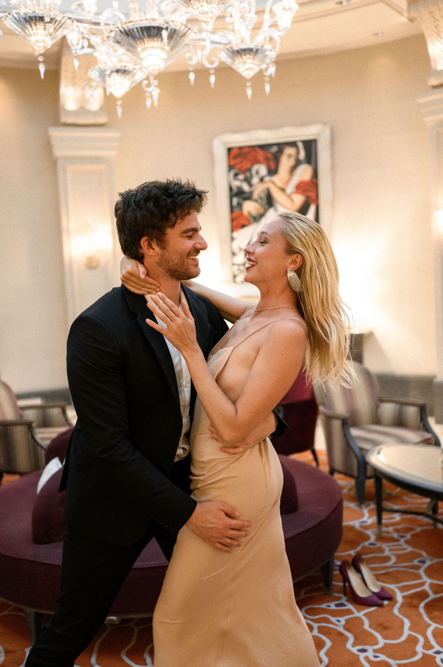 Elegant couple dancing and embracing in a luxurious hotel lobby under a crystal chandelier.